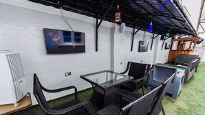 Un espacio de estar al aire libre techado, con mobiliario de mimbre, una mesa de vidrio y un televisor montado.