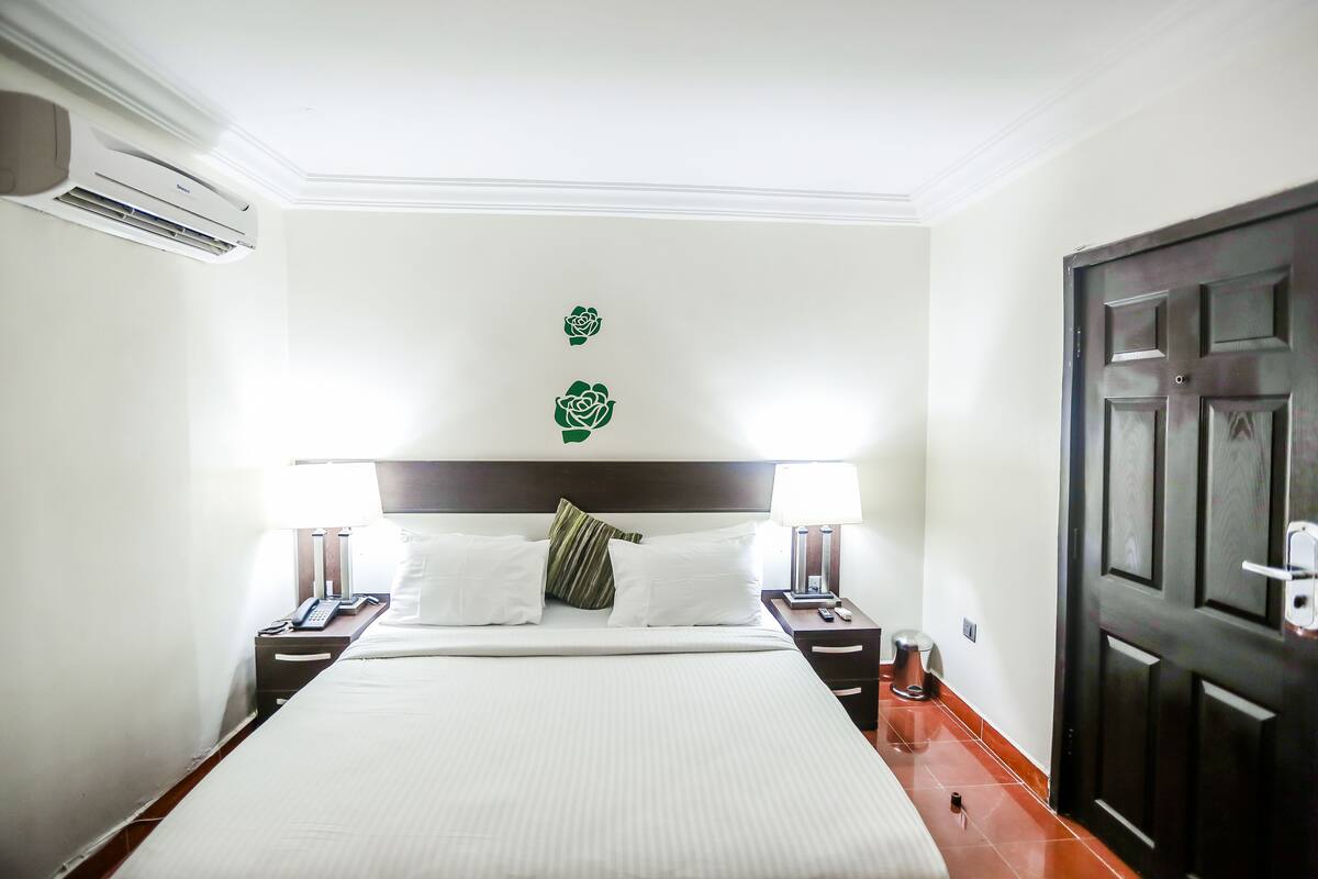A hotel room with a large bed, two bedside tables with lamps, a dark wooden door, and a wall-mounted air conditioner.
