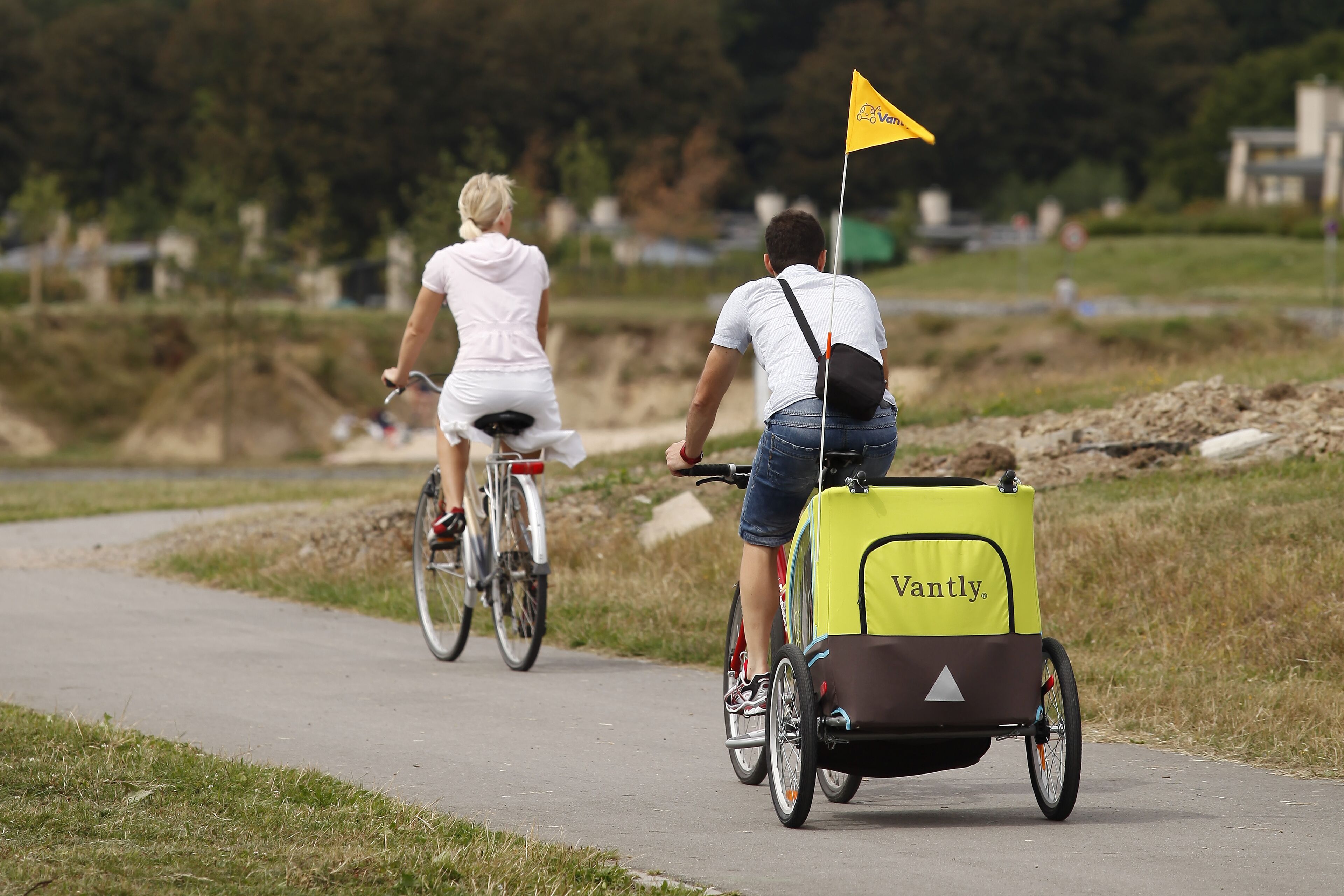 Dos personas montando en bicicleta por un camino pavimentado, con una bandera amarilla; una de ellas empuja un carro de reparto amarillo con la marca "Vantly".