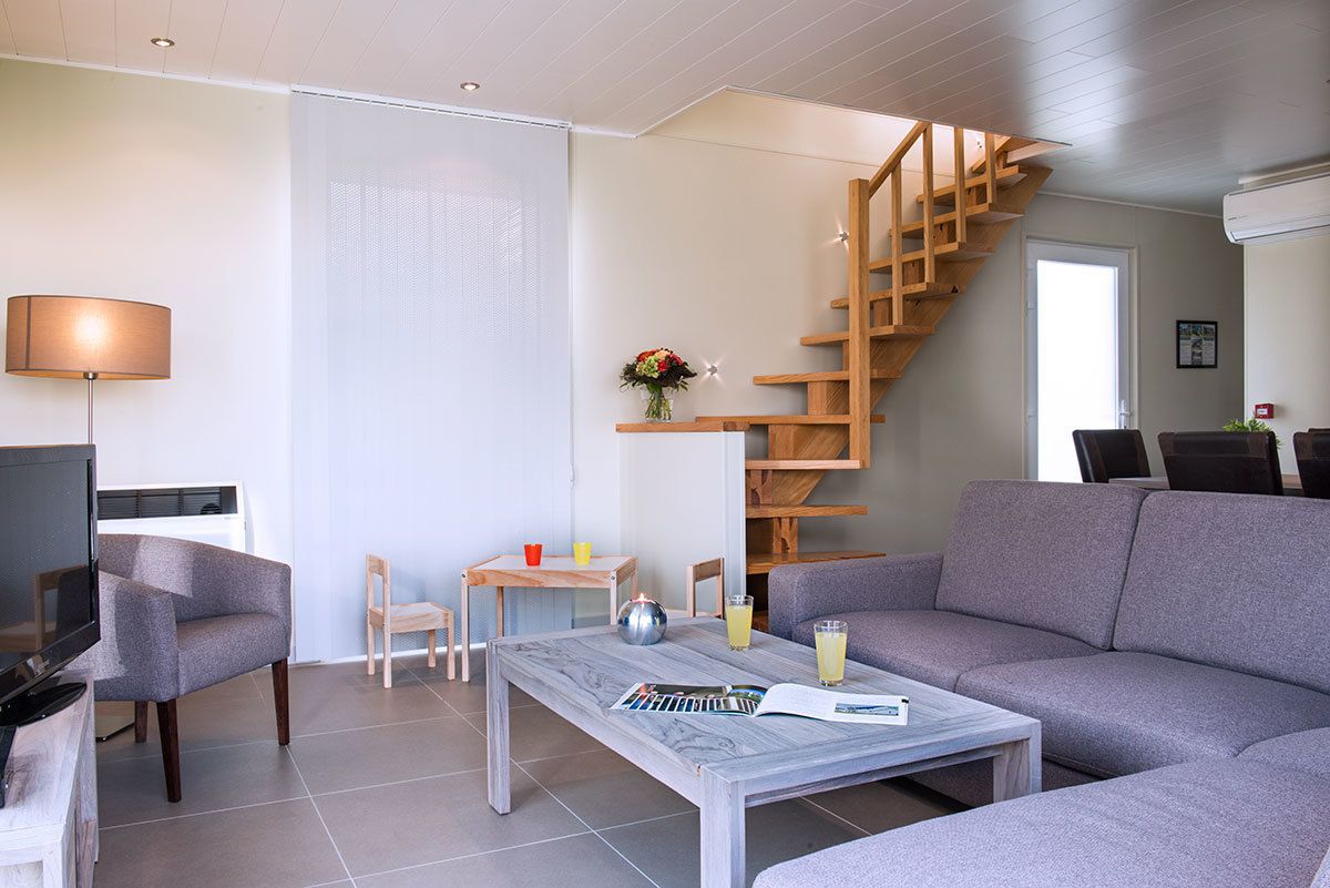 Una sala moderna con un sofá gris, una mesa de centro con revistas, un televisor, una escalera de madera y una mesita con un jarrón y bebidas.