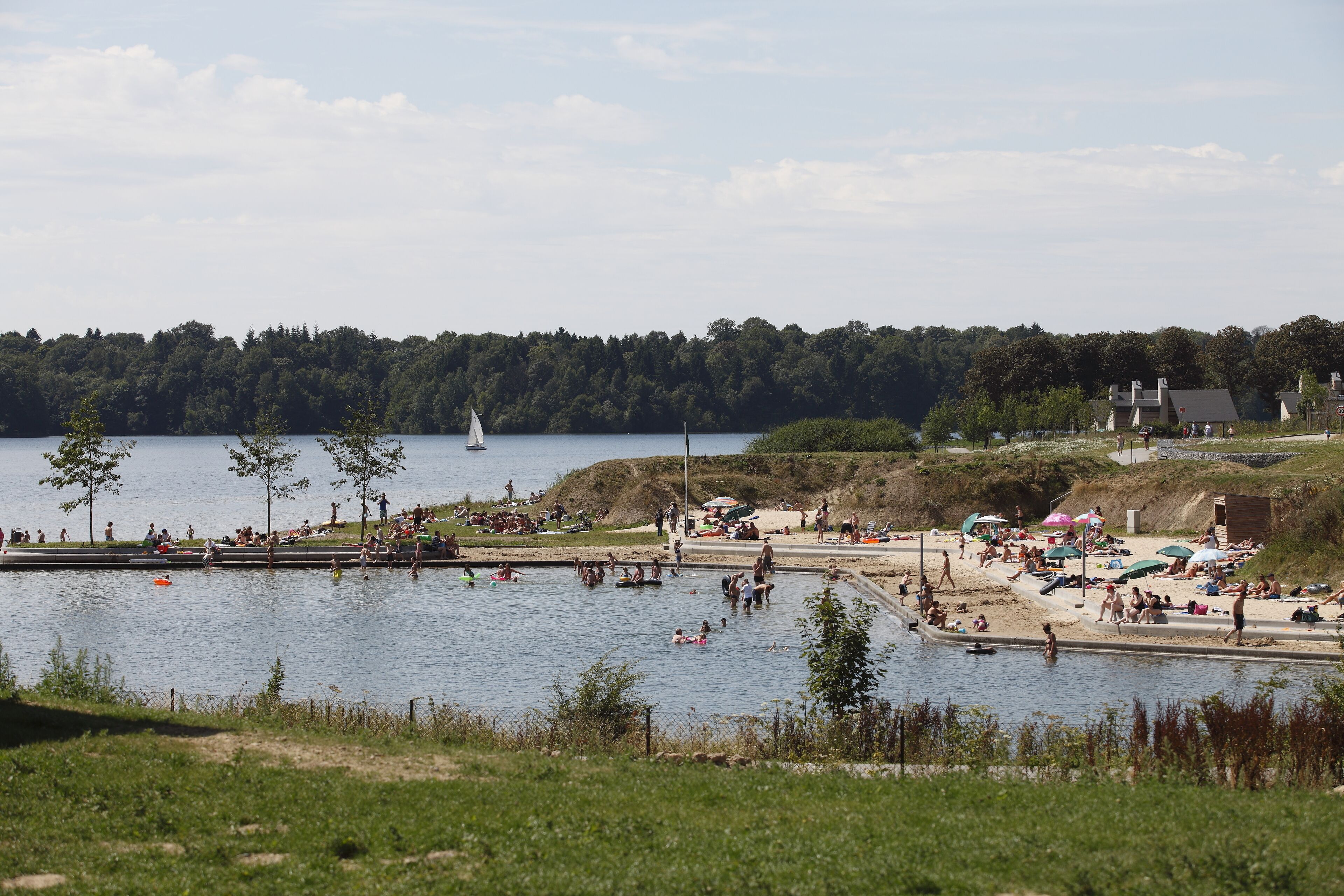 A lakeside area with a sandy beach, a small sailboat, and people engaging in various activities.