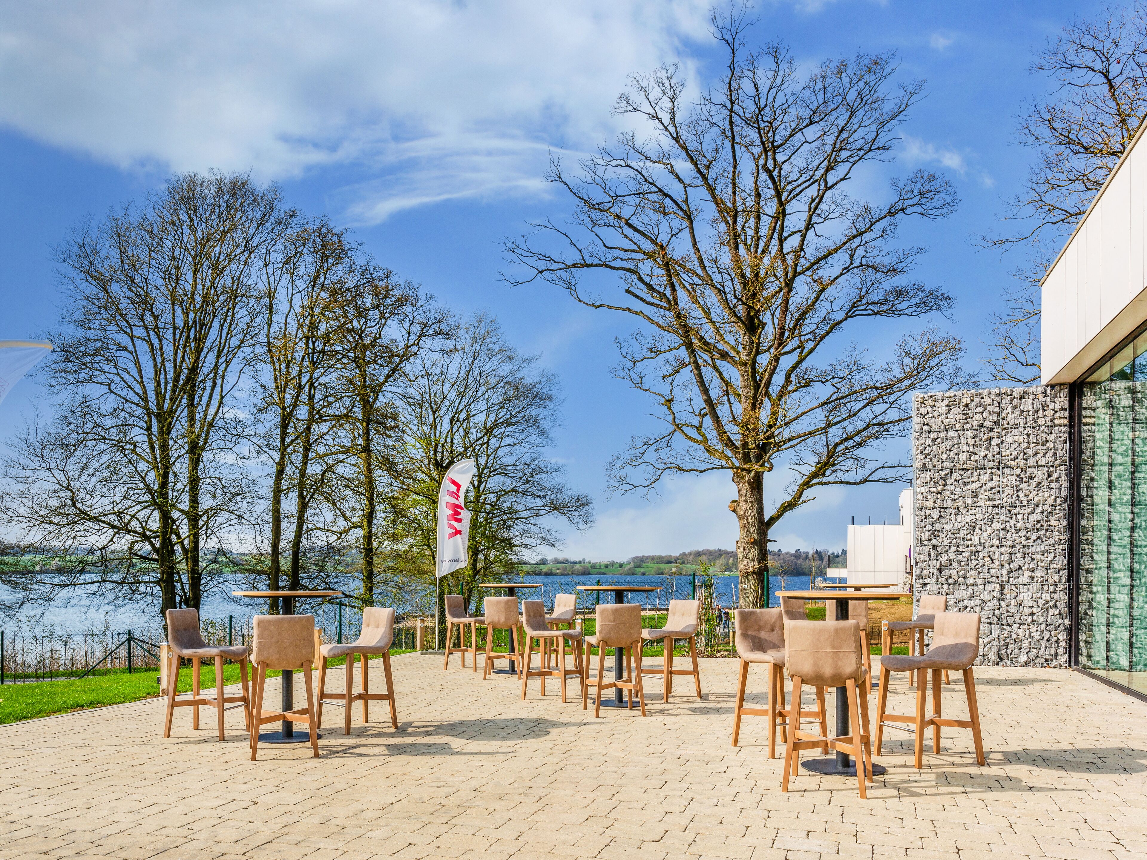 An outdoor seating area with chairs and tables, a flag, and trees against a backdrop of a building and a body of water.