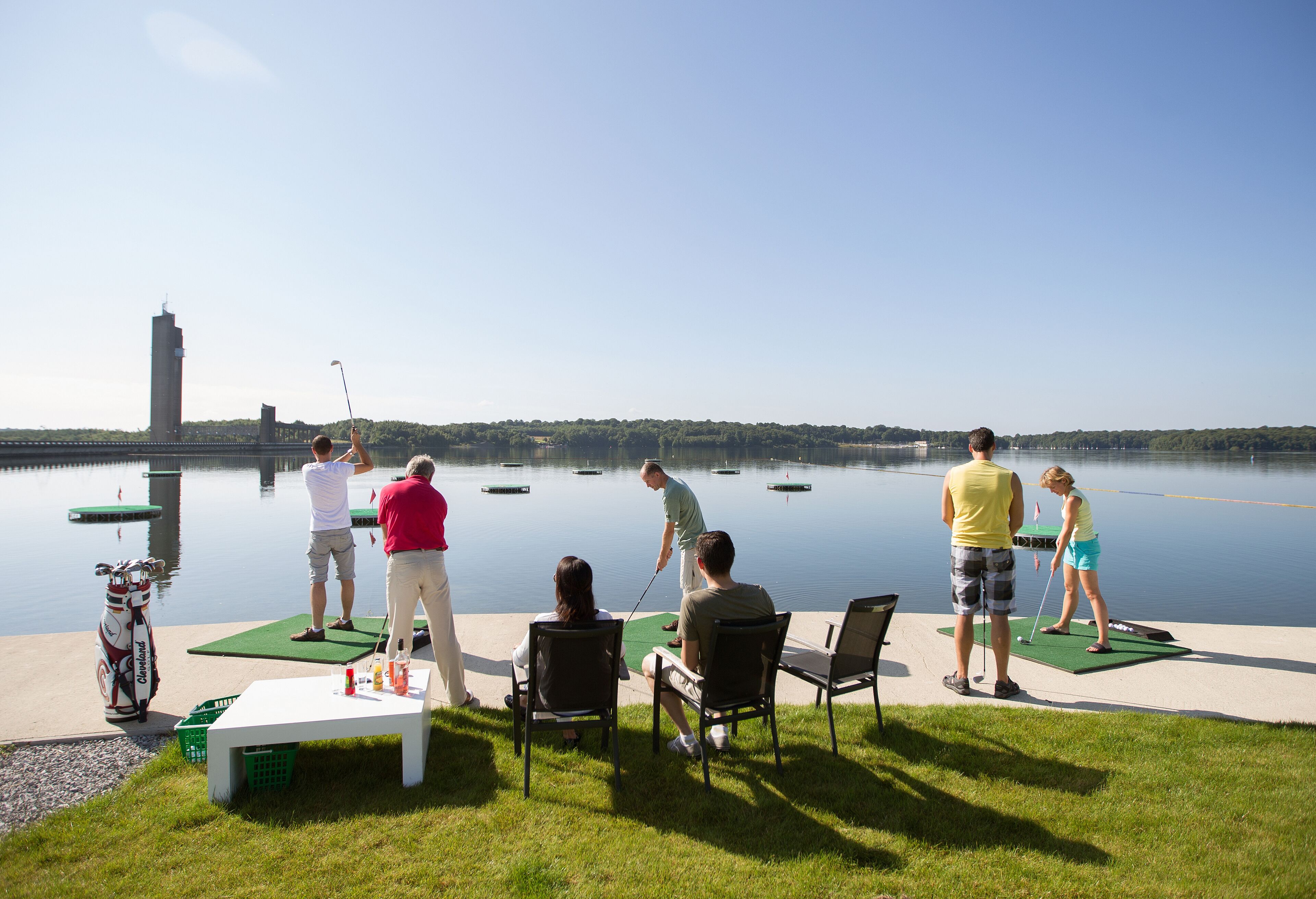 A group of people playing golf on a green near a body of water with a building in the background.