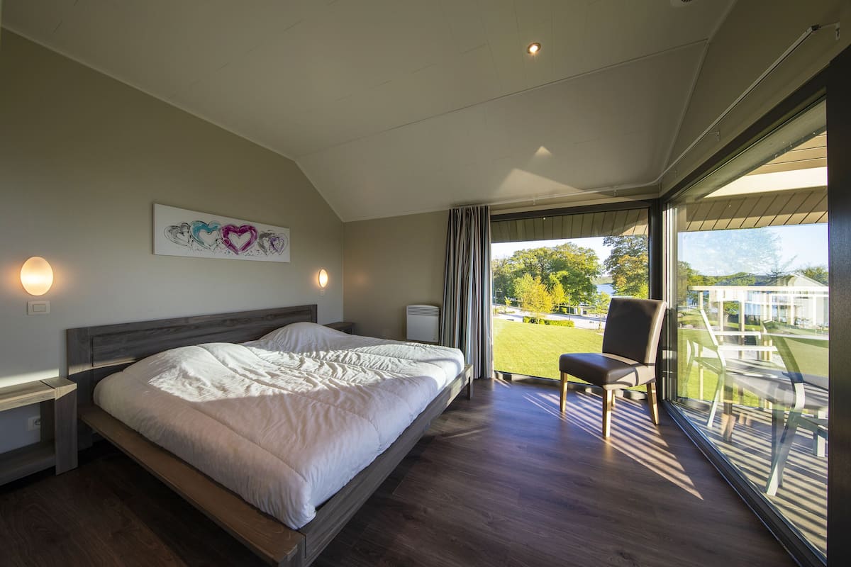 A modern bedroom with a large bed, a chair, and a view of the outdoors through large windows.