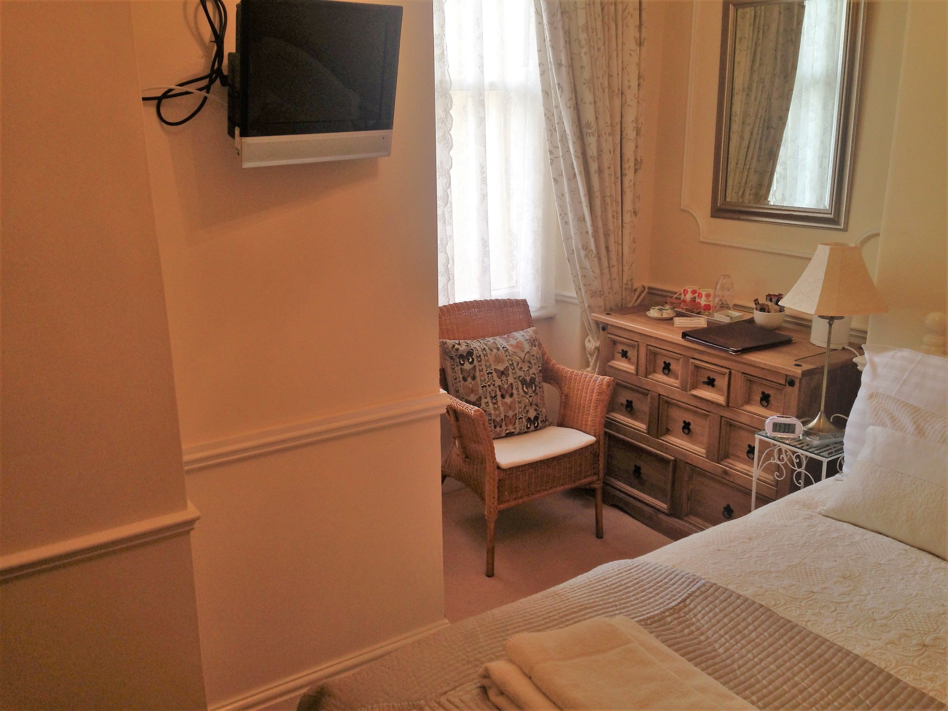 Un dormitorio con cama, una silla, cómoda, televisor montado en la pared y ventana con cortinas.