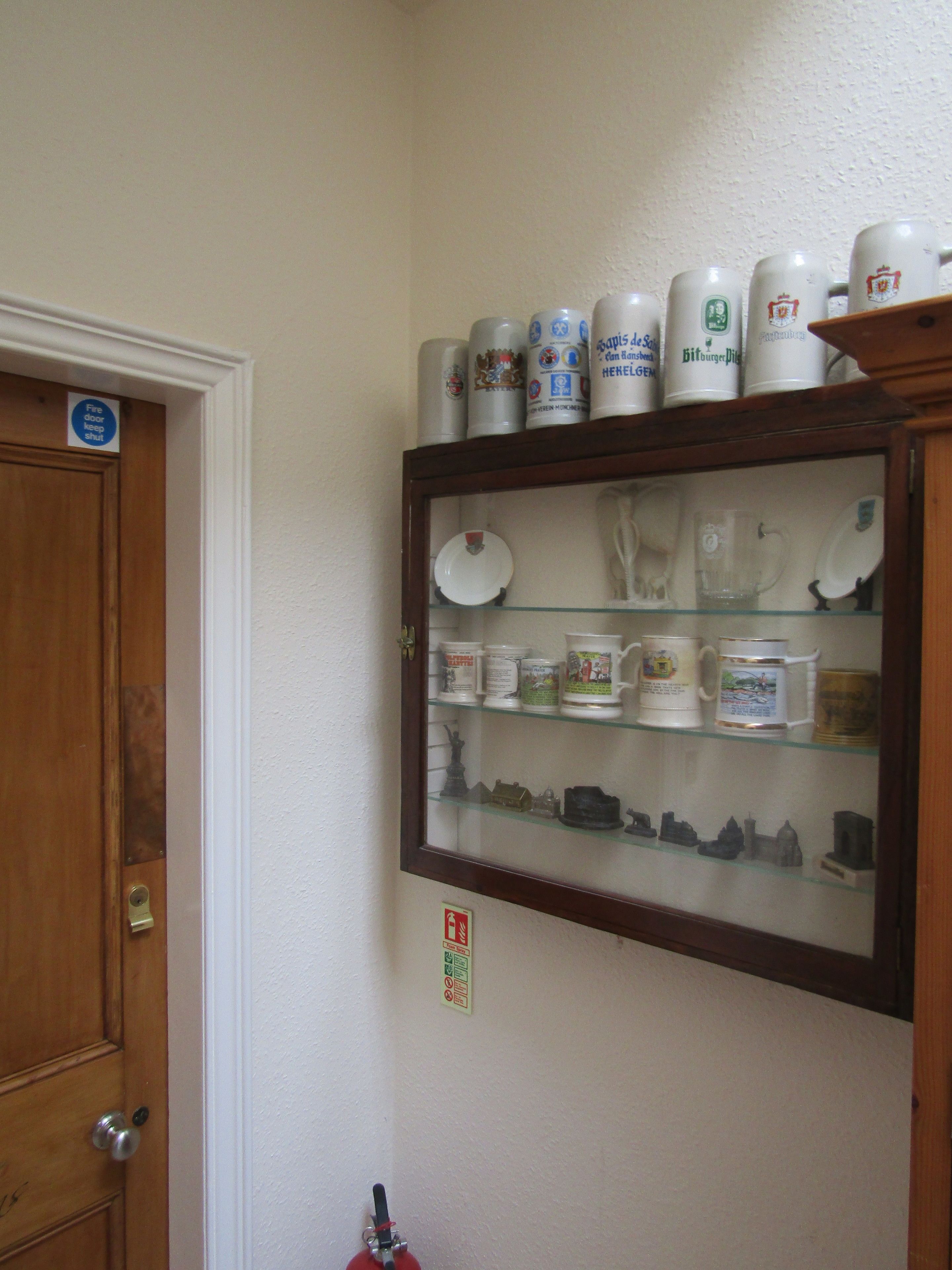 Eine Holztür mit Messingklinke, eine Vitrine mit verschiedenen Keramikgegenständen und ein Brandschutznachweis an der Wand.