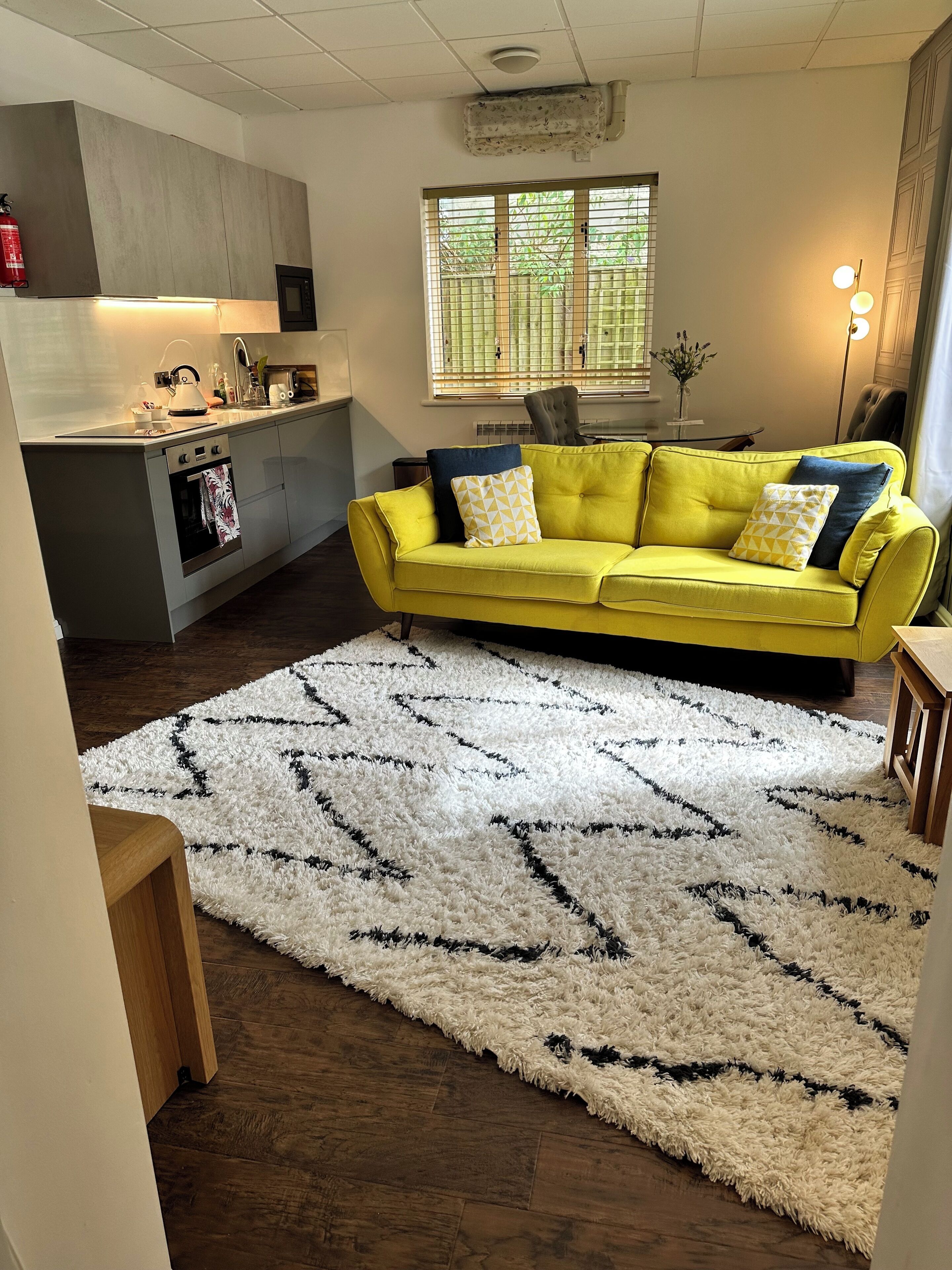 A modern living room with a yellow sofa, a white shag rug, and a kitchen in the background.
