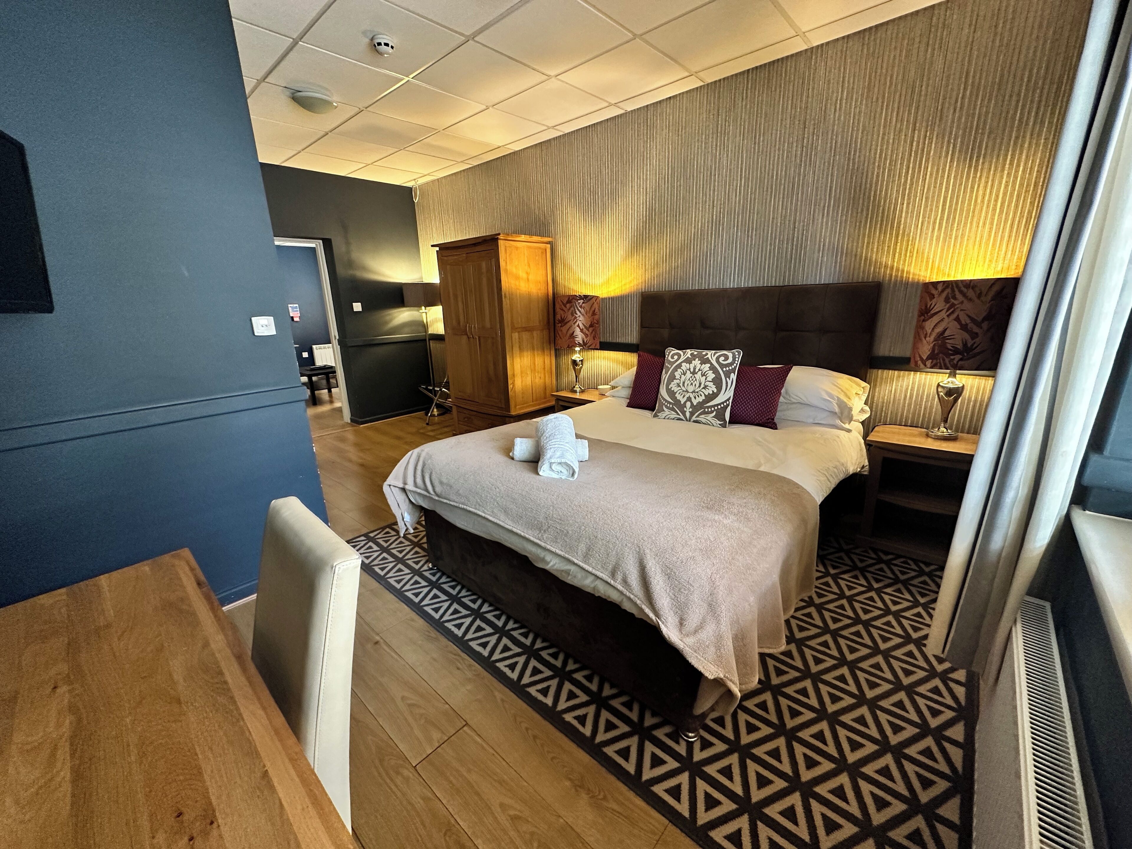 A hotel room with a large bed, a desk, a wardrobe, and a small kitchenette.