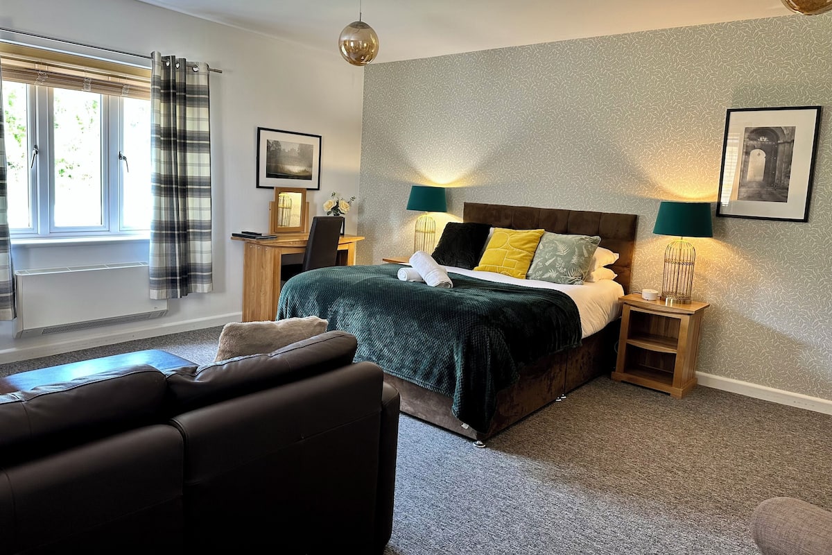A bedroom with a bed, two bedside tables, a desk, a chair, a sofa, and a window with curtains.