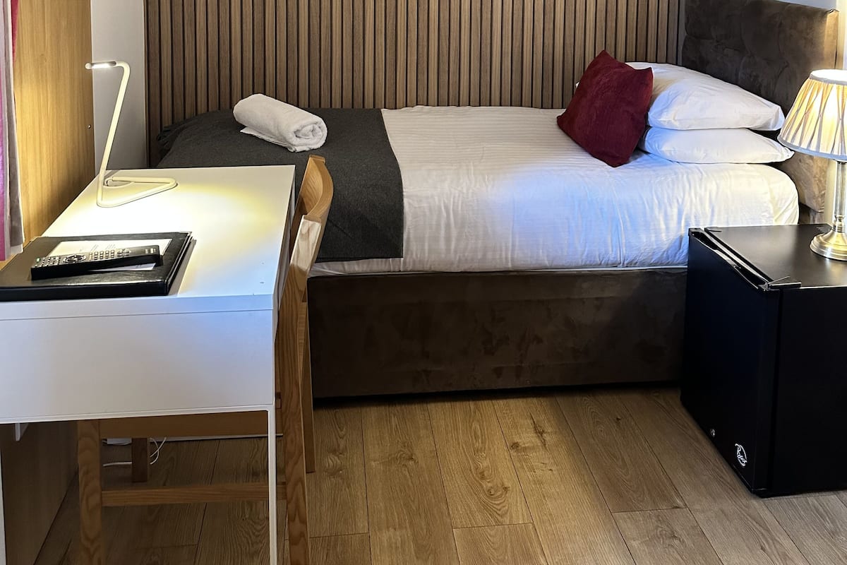 A hotel room with a wooden slat headboard, a bed with white bedding and a red pillow, a bedside table with a lamp, and a desk with a computer.