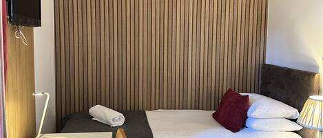 Een hotelkamer met een houten lattenkap, een bed met wit beddengoed en een rode kussen, een nachtkastje met een lamp, en een bureau met een computer.
