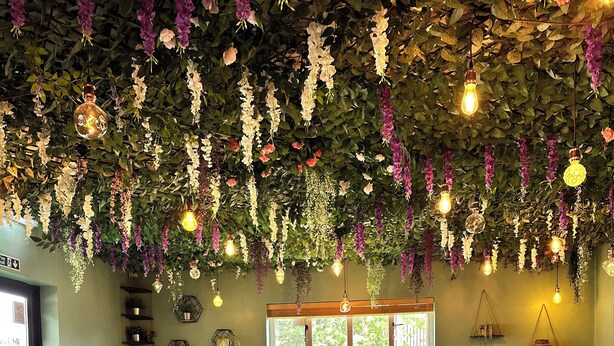 A restaurant interior with wooden tables and chairs, hanging floral decorations, and a view of the outdoors through large windows. - The Churchill Court (Witney)