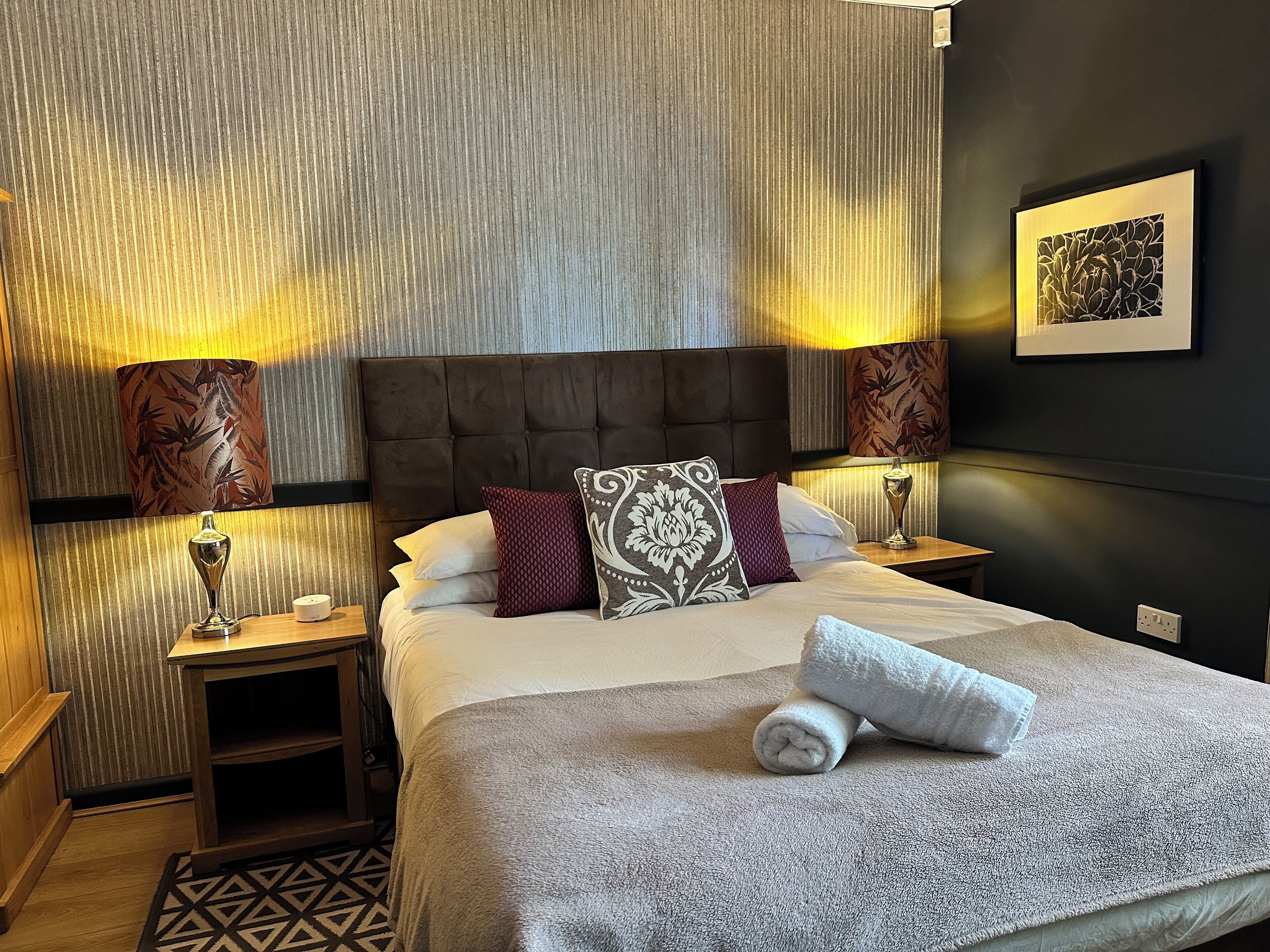 A neatly made bed with a grey blanket and a rolled towel, flanked by two bedside lamps with patterned lampshades, a small wooden nightstand, and a framed artwork on the wall.