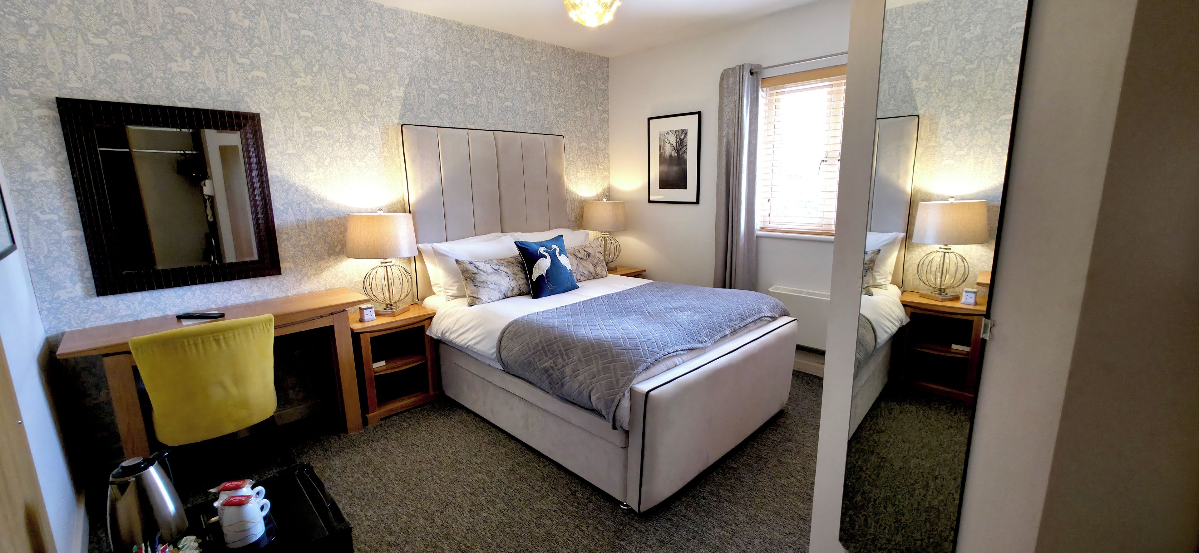 A hotel room with a bed, bedside tables, a desk, a chair, a mirror, and a window with curtains.