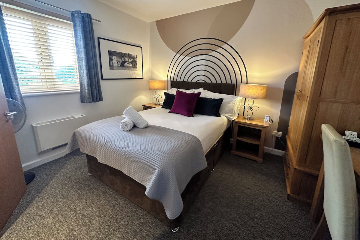 A hotel room with a large bed, a wooden wardrobe, a bedside table with a lamp, a picture on the wall, and a window with curtains.