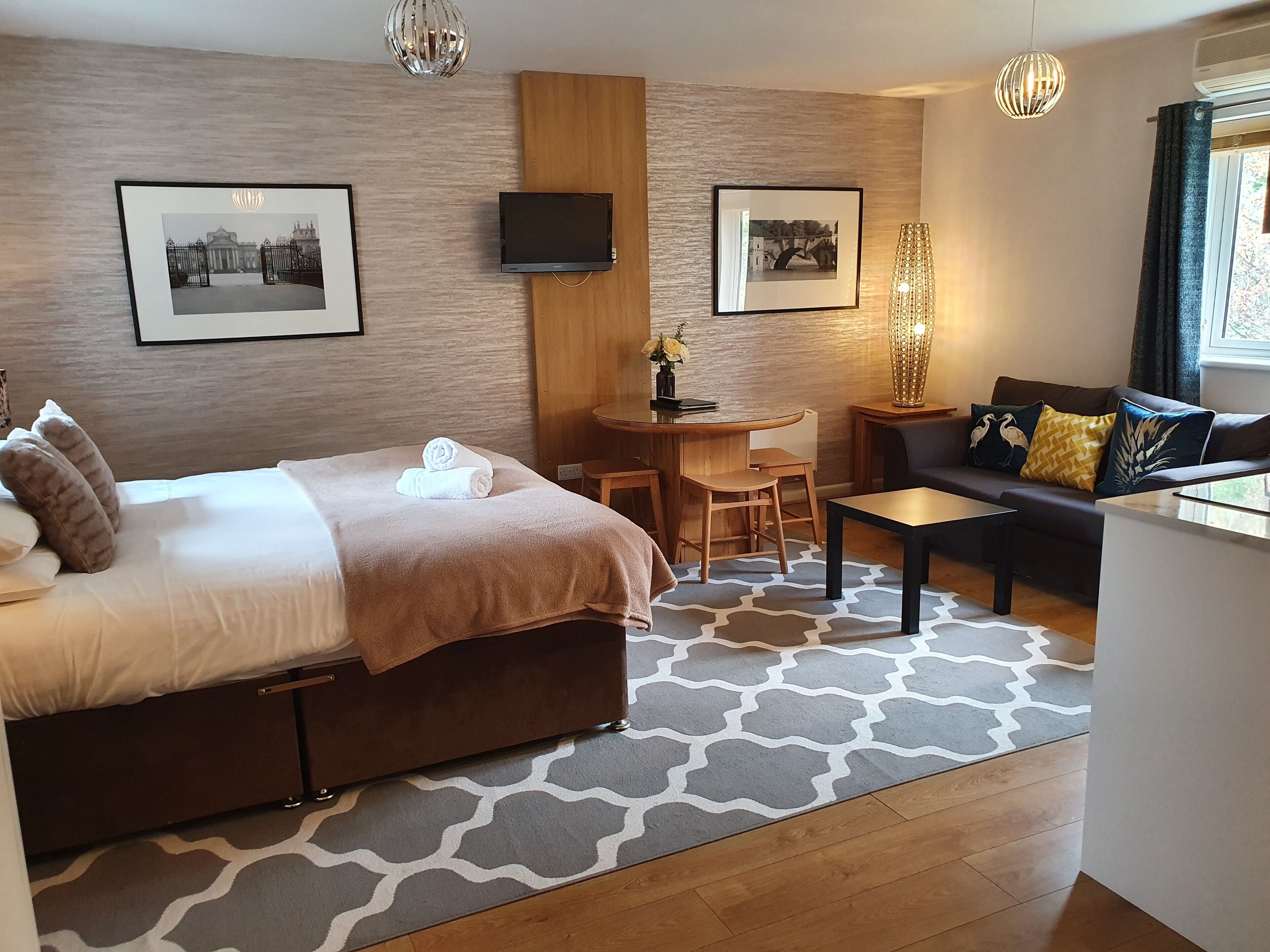 A modern hotel room with a bed, a sofa, a small table, and a TV mounted on the wall.