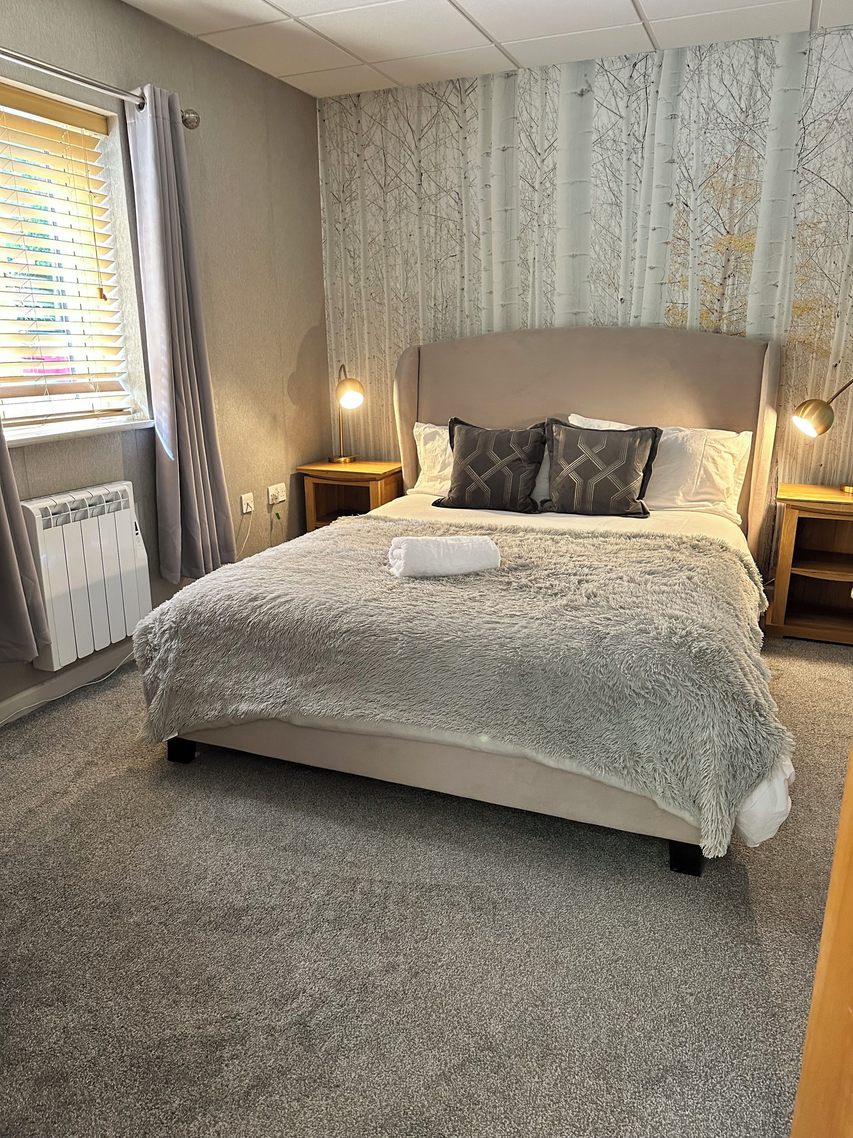 Un lit bien fait, recouvert d’une couverture blanche et moelleuse, deux lampes de chevet, un radiateur et une fenêtre avec des stores.