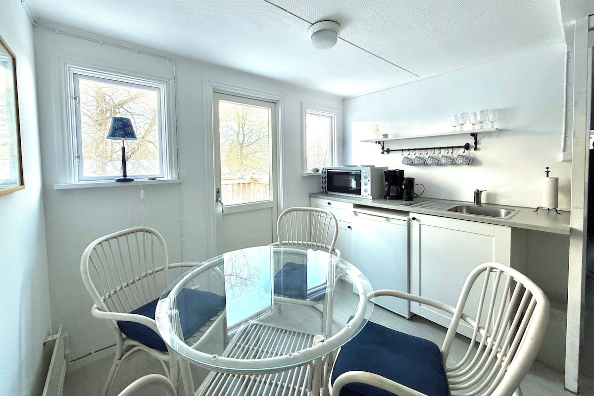 A compact kitchen with a microwave, coffee maker, and a small sink. There is a dining table with chairs and a window with a view of trees.