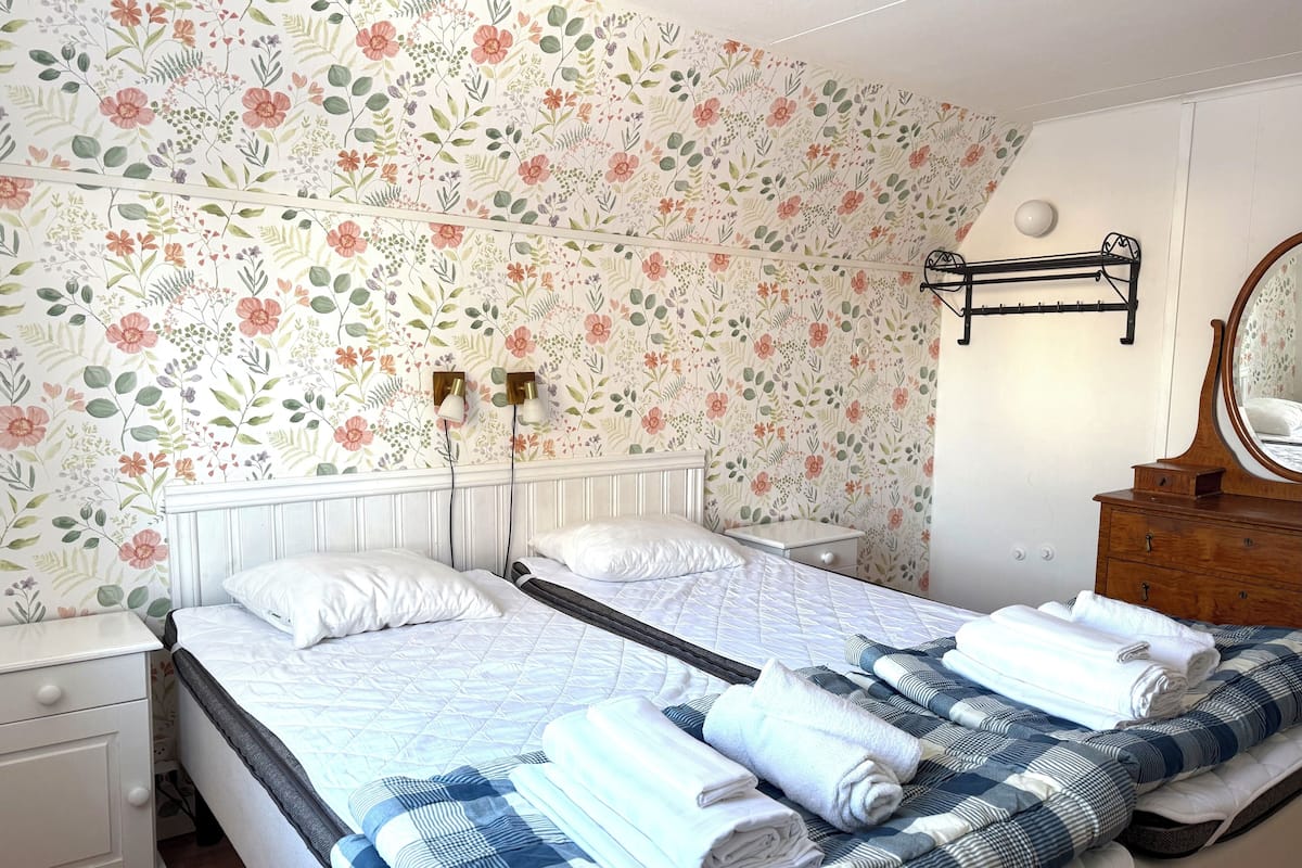 A double bed with a quilted bedspread, two bedside tables with lamps, a wooden dresser with a mirror, and floral wallpaper.