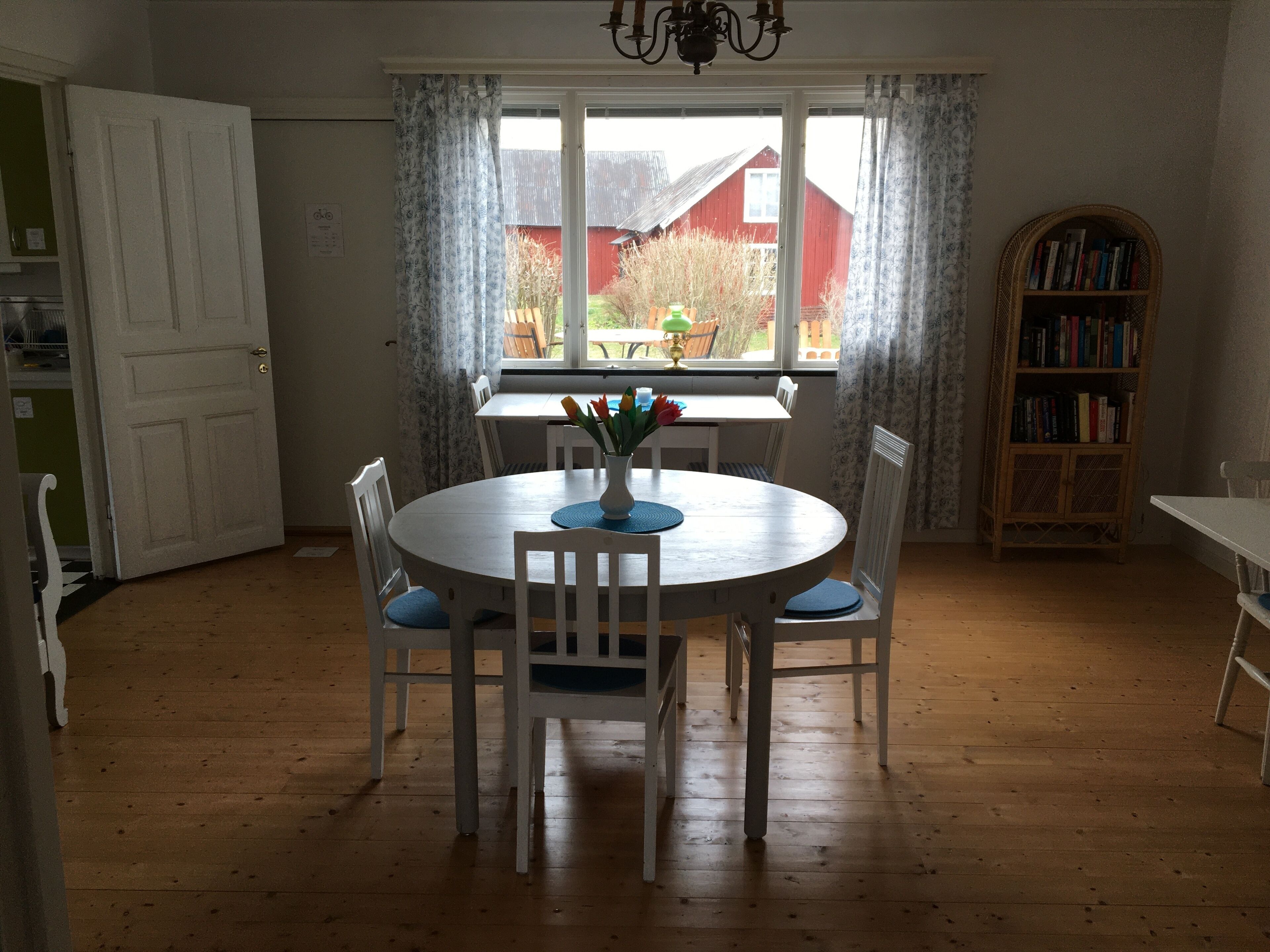 A dining area with a round table, white chairs, and a vase with flowers. There is a window with curtains, a bookshelf, and a door.