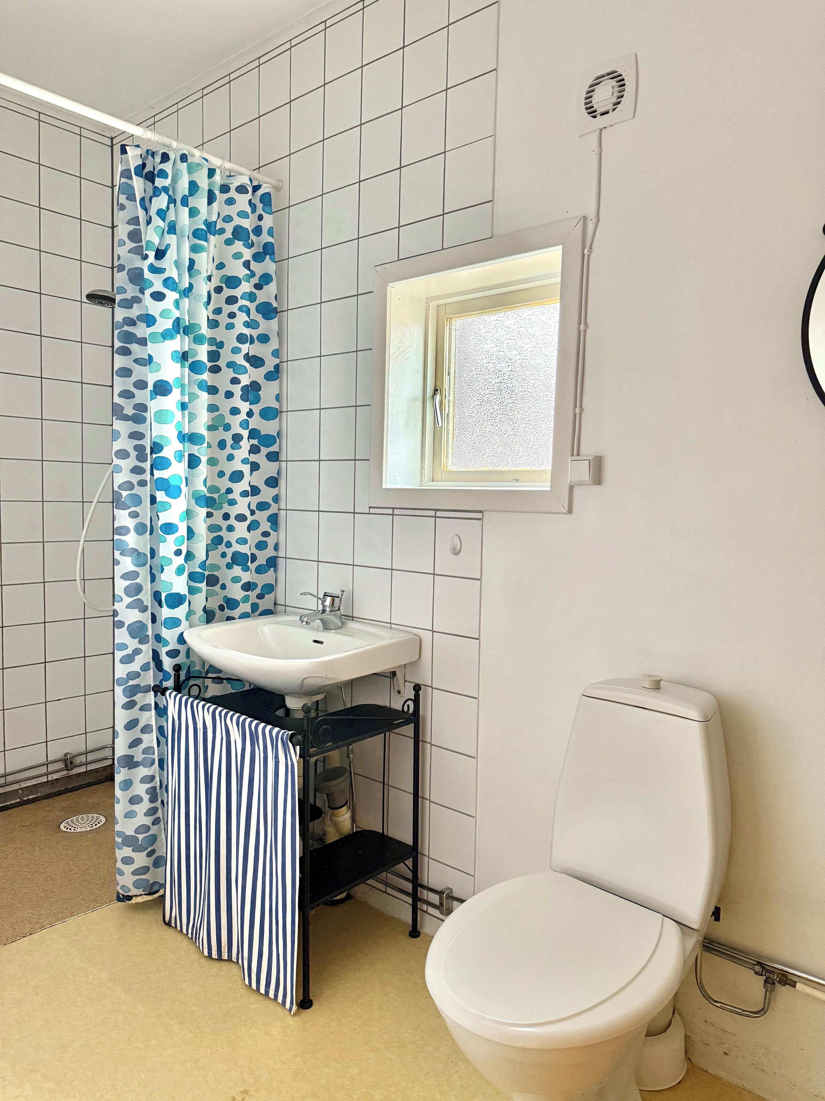 A bathroom with a white toilet, a white sink, a shower with a blue polka dot curtain, and a small window above the sink.