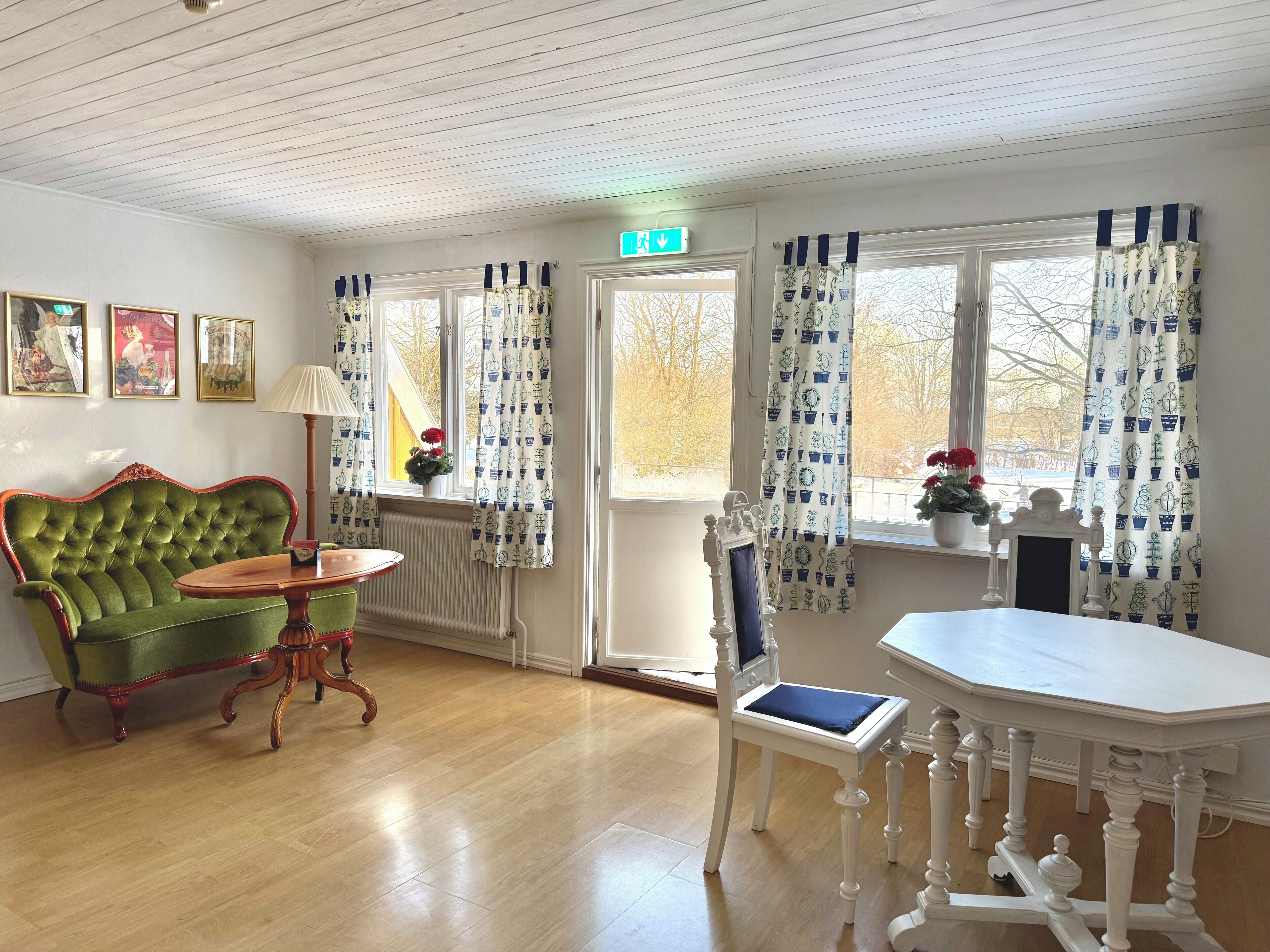 Una habitación con un sofá tapizado en verde, una mesa de madera, una silla blanca con un cojín azul y una mesa blanca con patas decoradas.