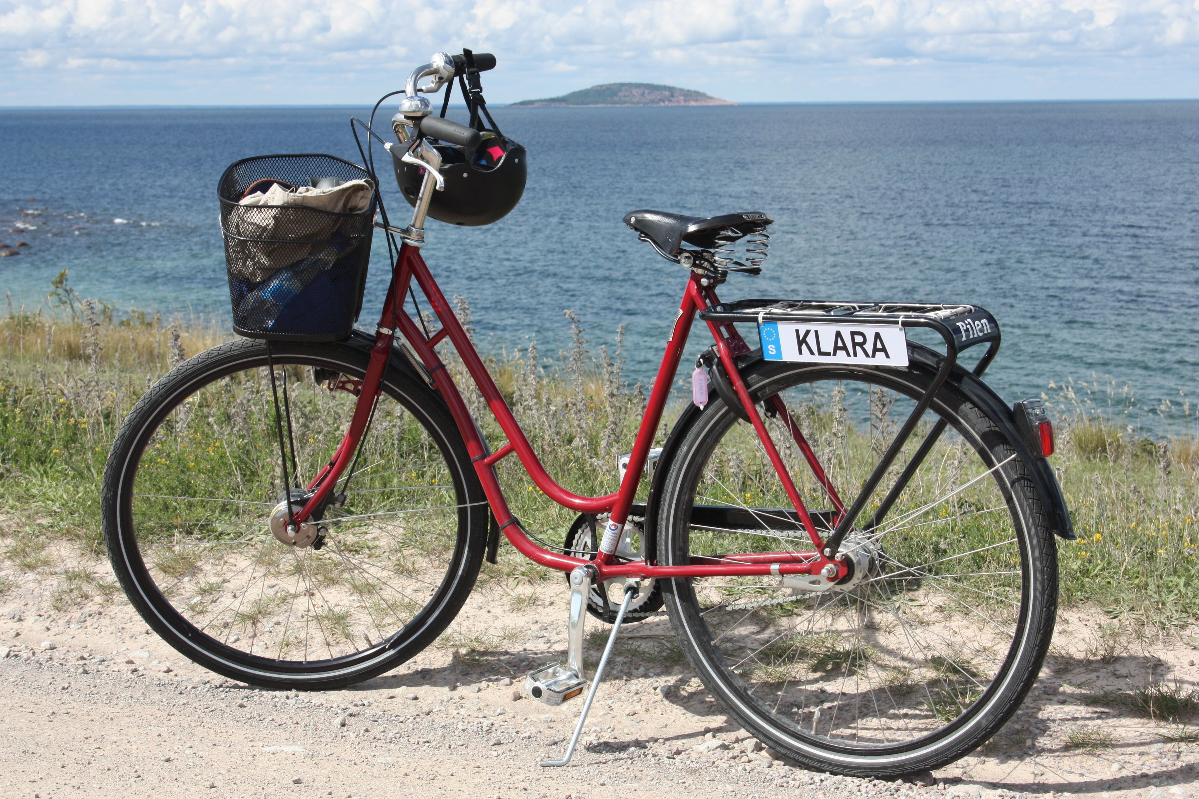 A red bicycle with a basket and a sign reading "KLARA" parked on a dirt path by the sea.