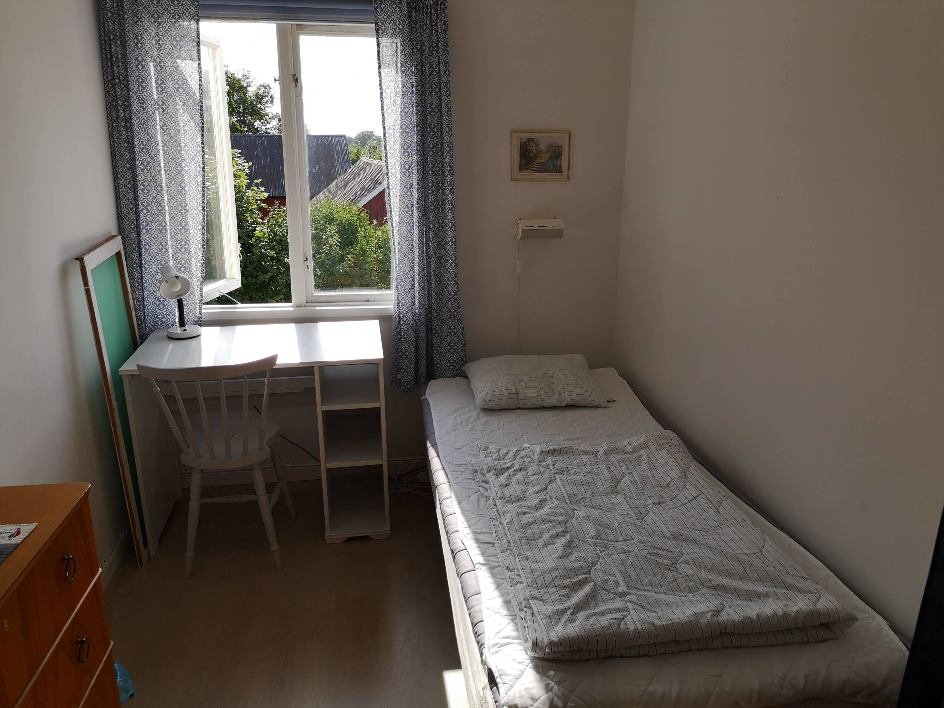 Een kleine, net kamer met een enkel bed, een bureau met stoel en een raam met gordijnen met dessin.