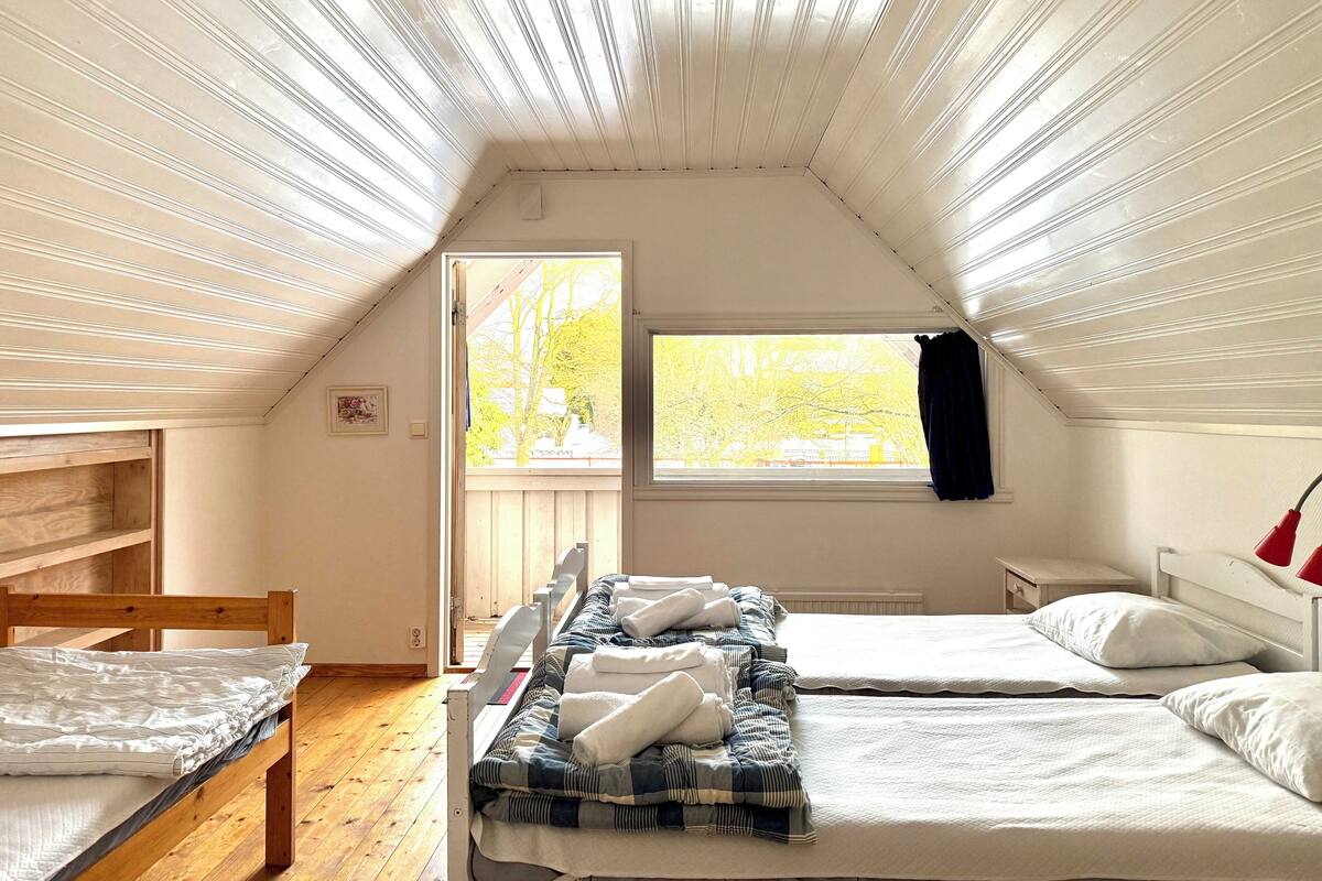 A cozy attic bedroom with two beds, a window with a view of trees, a wooden chair, and a small table.