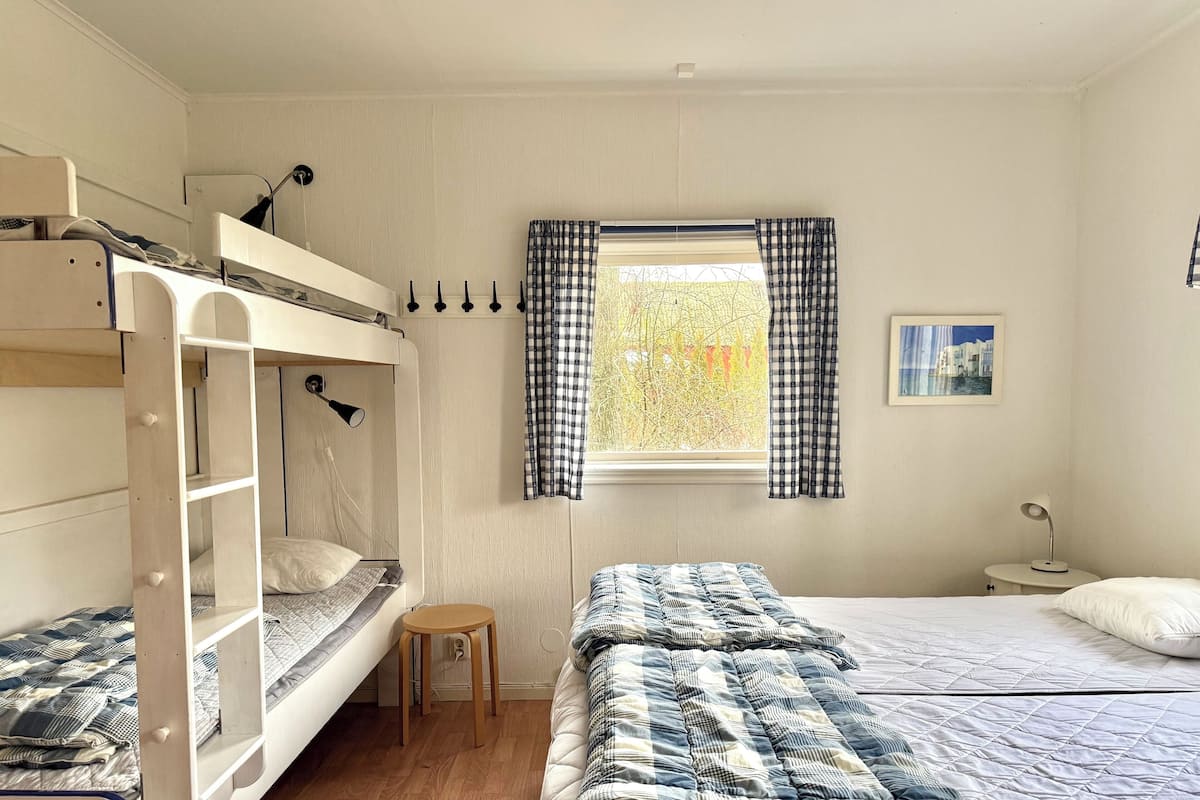 A dorm-style room with bunk beds, a small stool, a window with checkered curtains, and a bedside table with a lamp.