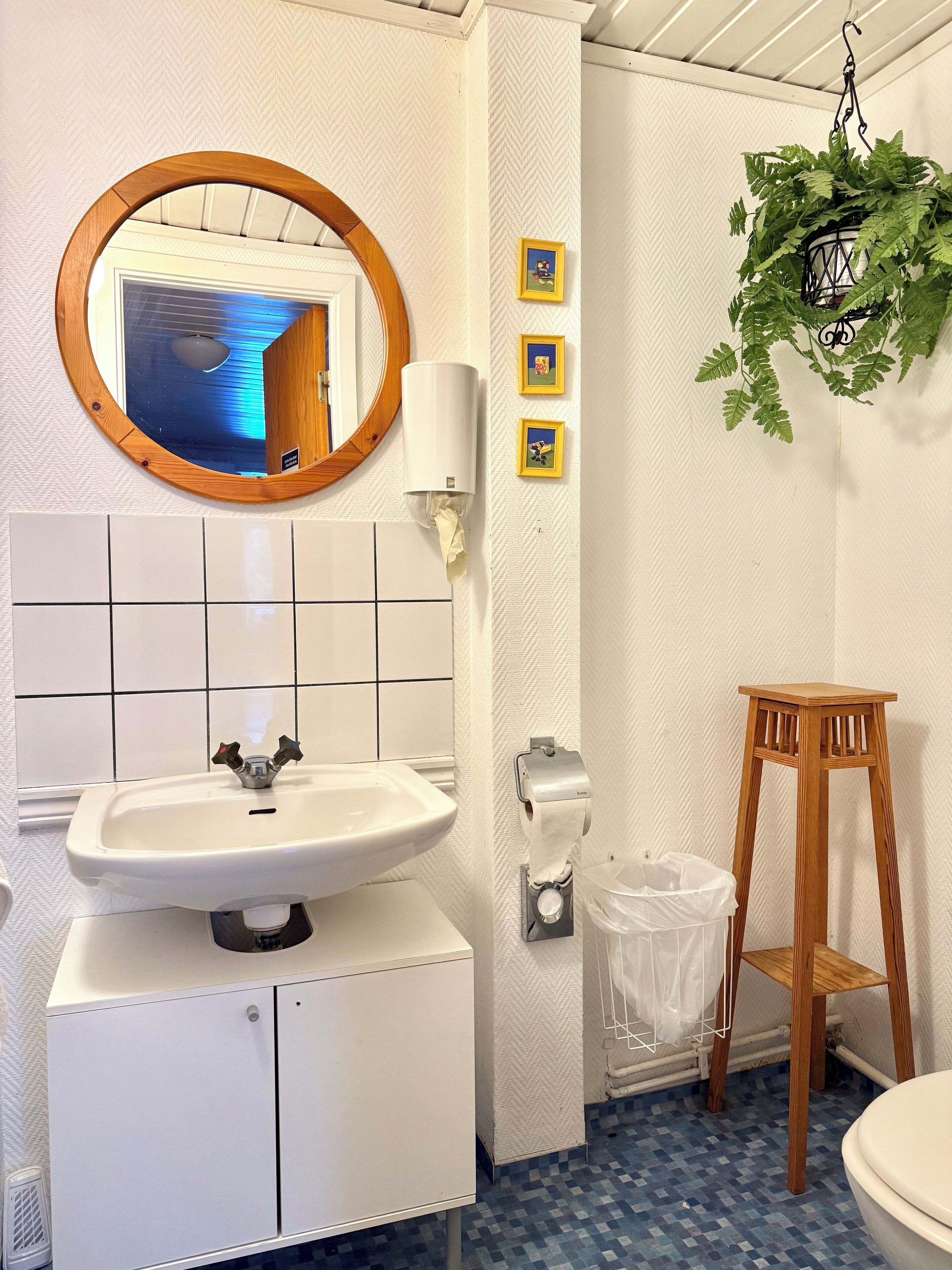 A bathroom with a white sink, mirror, and tiled walls. There is a wooden stool and a trash bin.