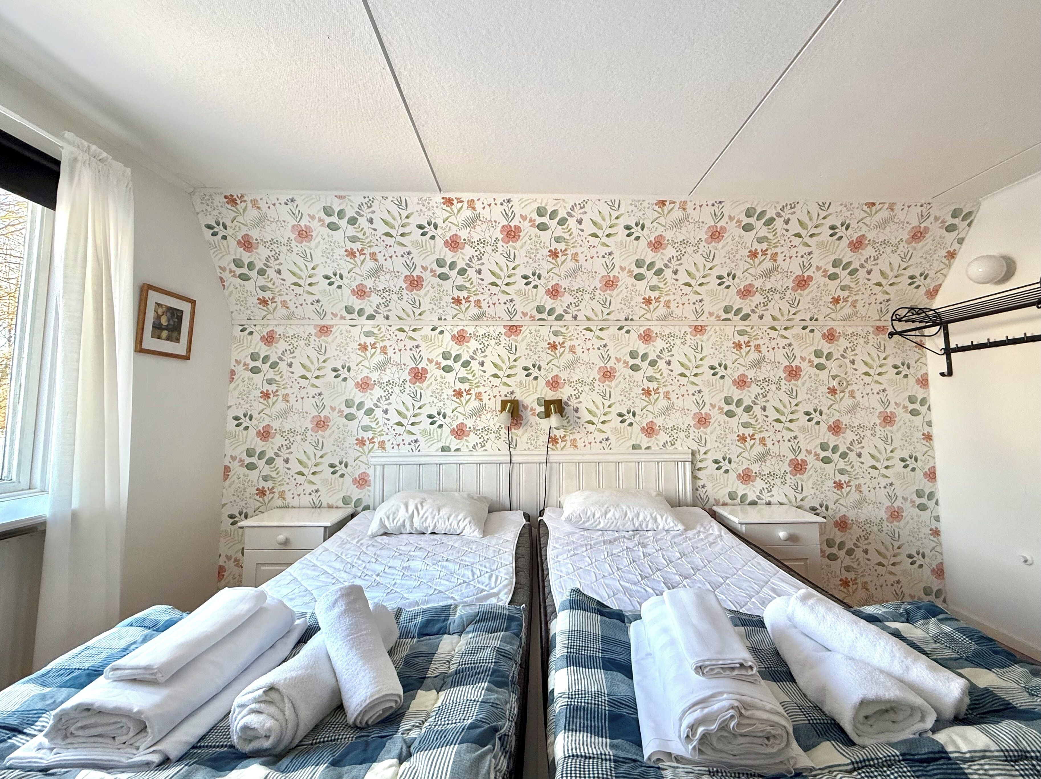 Habitación doble con papel tapiz floral, dos camas con toallas enrolladas, una mesita de noche, una lámpara, un cuadro en la pared y una ventana con cortinas.