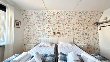 Habitación doble con papel tapiz floral, dos camas con toallas enrolladas, una mesita de noche, una lámpara, un cuadro en la pared y una ventana con cortinas.