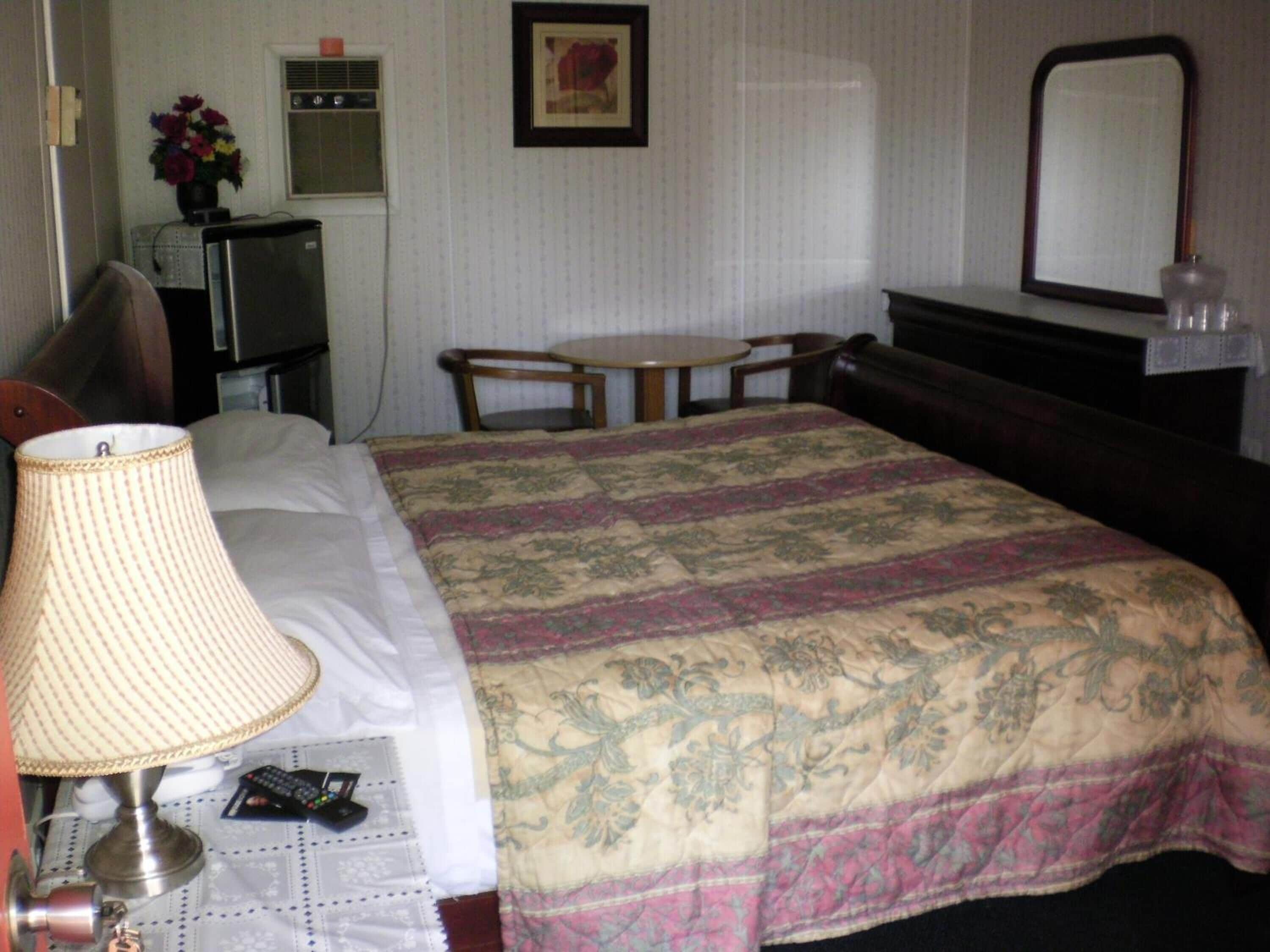 Une chambre d’hôtel comprenant un lit, une table de chevet avec une lampe, une petite table avec des chaises, une commode, un miroir et un radiateur fixé au mur.