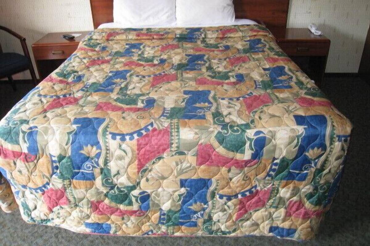 A bed with a colorful quilt, a wooden headboard, a bedside table, and a chair.