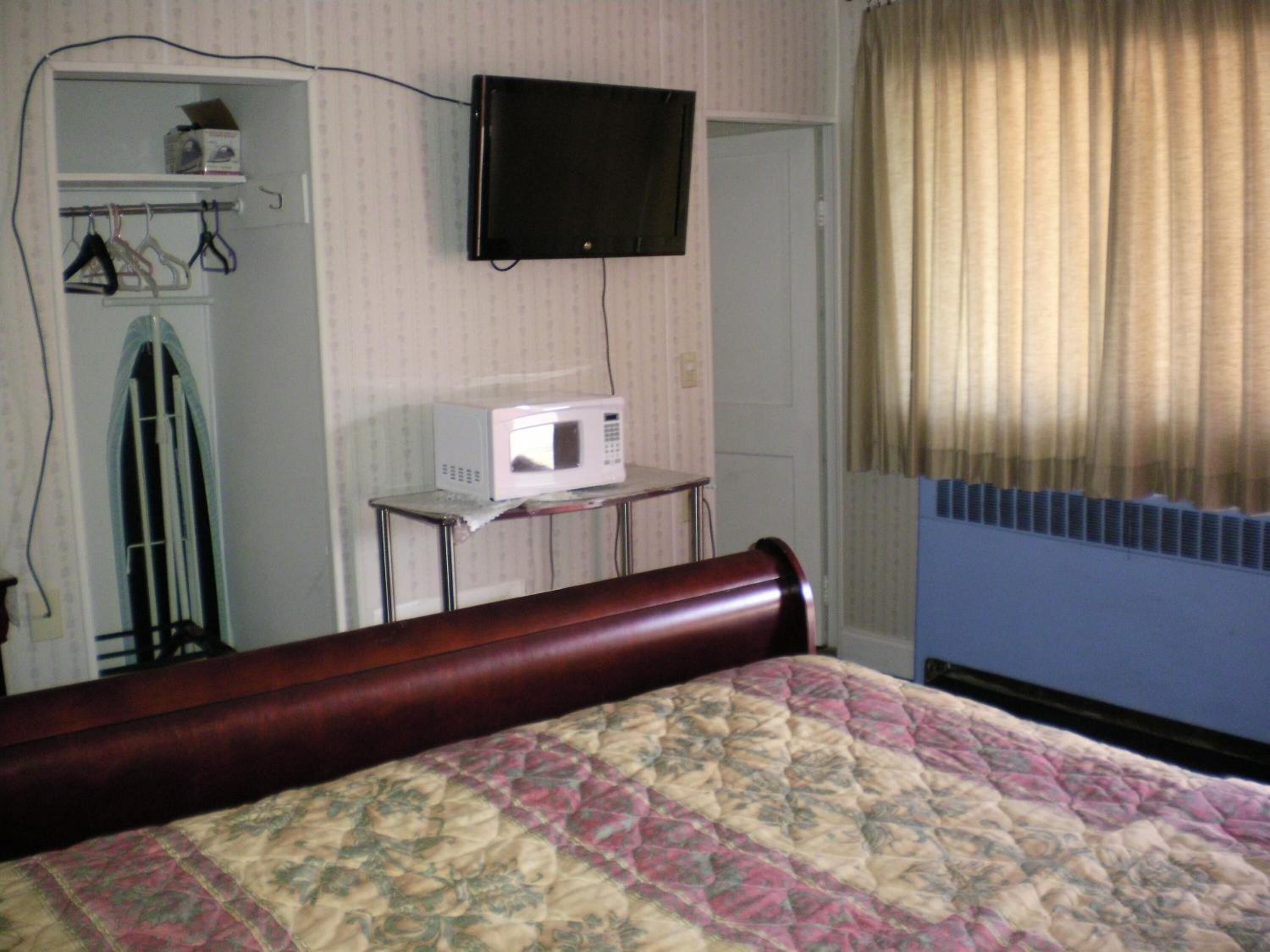 A bedroom with a bed, a television mounted on the wall, a microwave on a small table, and a window with curtains.