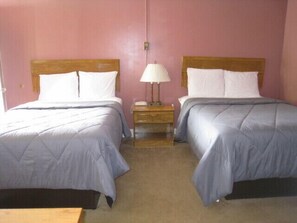 A room with two beds, each with a wooden headboard and white bedding. There is a nightstand with a lamp between the beds.