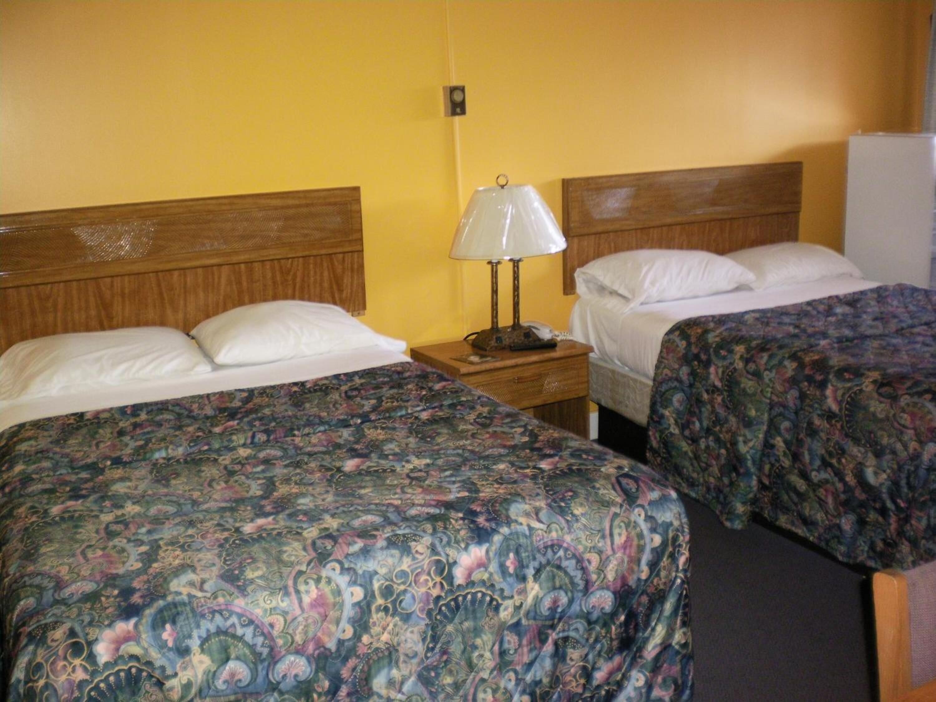 A hotel room with two beds, each with a wooden headboard, a nightstand with a lamp, and a white refrigerator.
