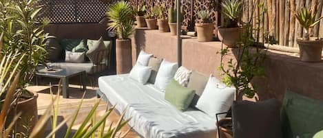 An outdoor seating area with a sofa, chairs, and a table, surrounded by potted plants and a shaded canopy.