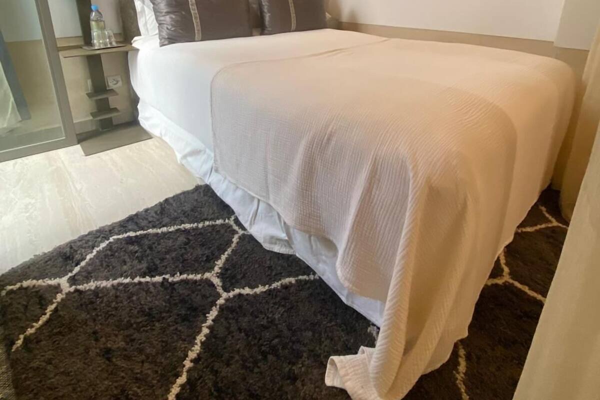 A neatly made bed with white linens, a dark patterned rug, and two wall-mounted lamps.