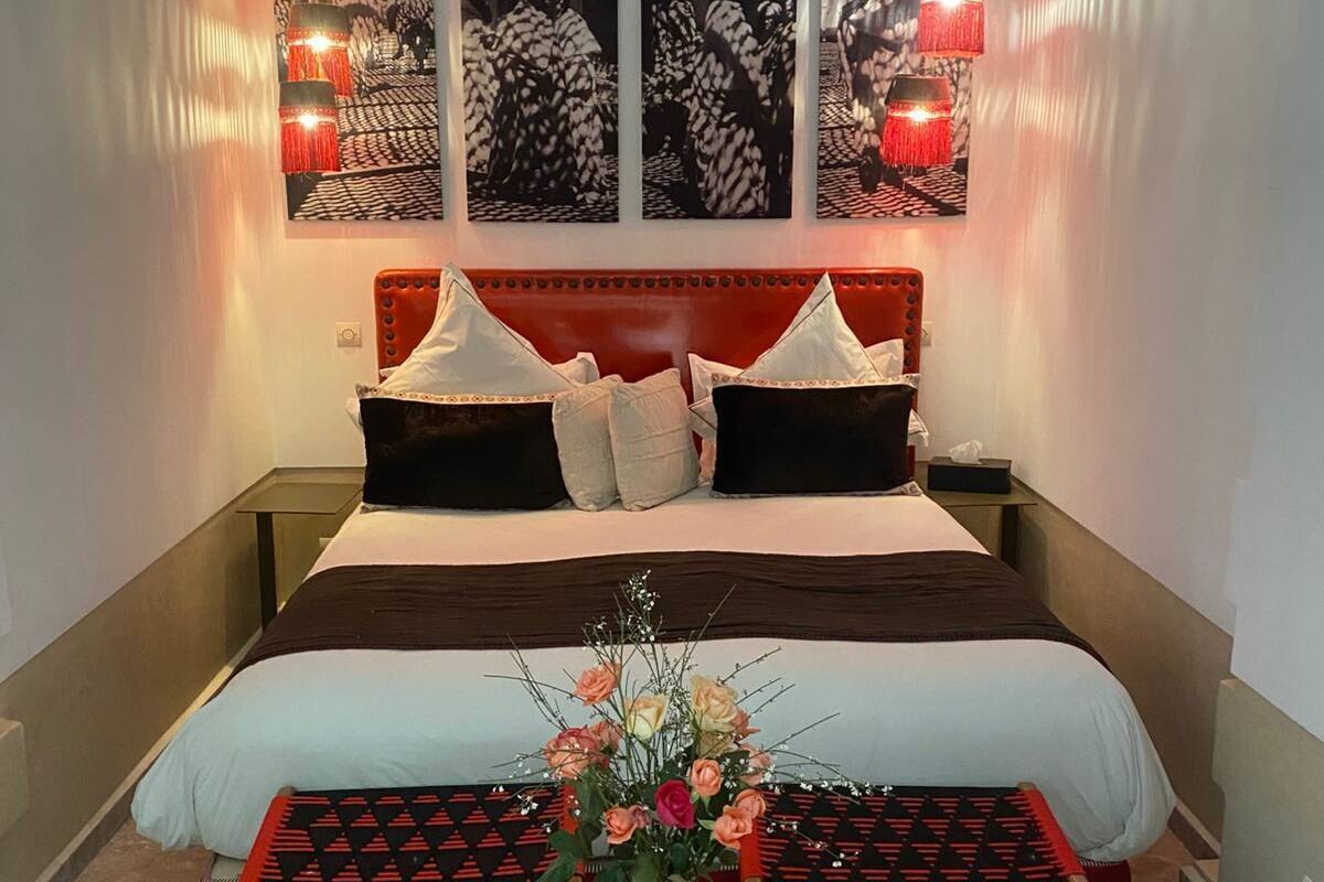A bedroom with a red upholstered headboard, a large bed with multiple pillows, a small round table with a vase of flowers, and three wall-mounted lamps.