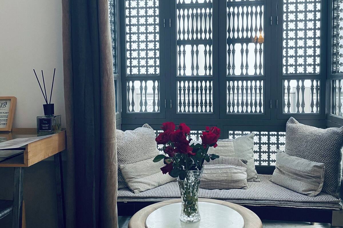 A room with a large window, a round table with a vase of flowers, and a sofa with cushions.
