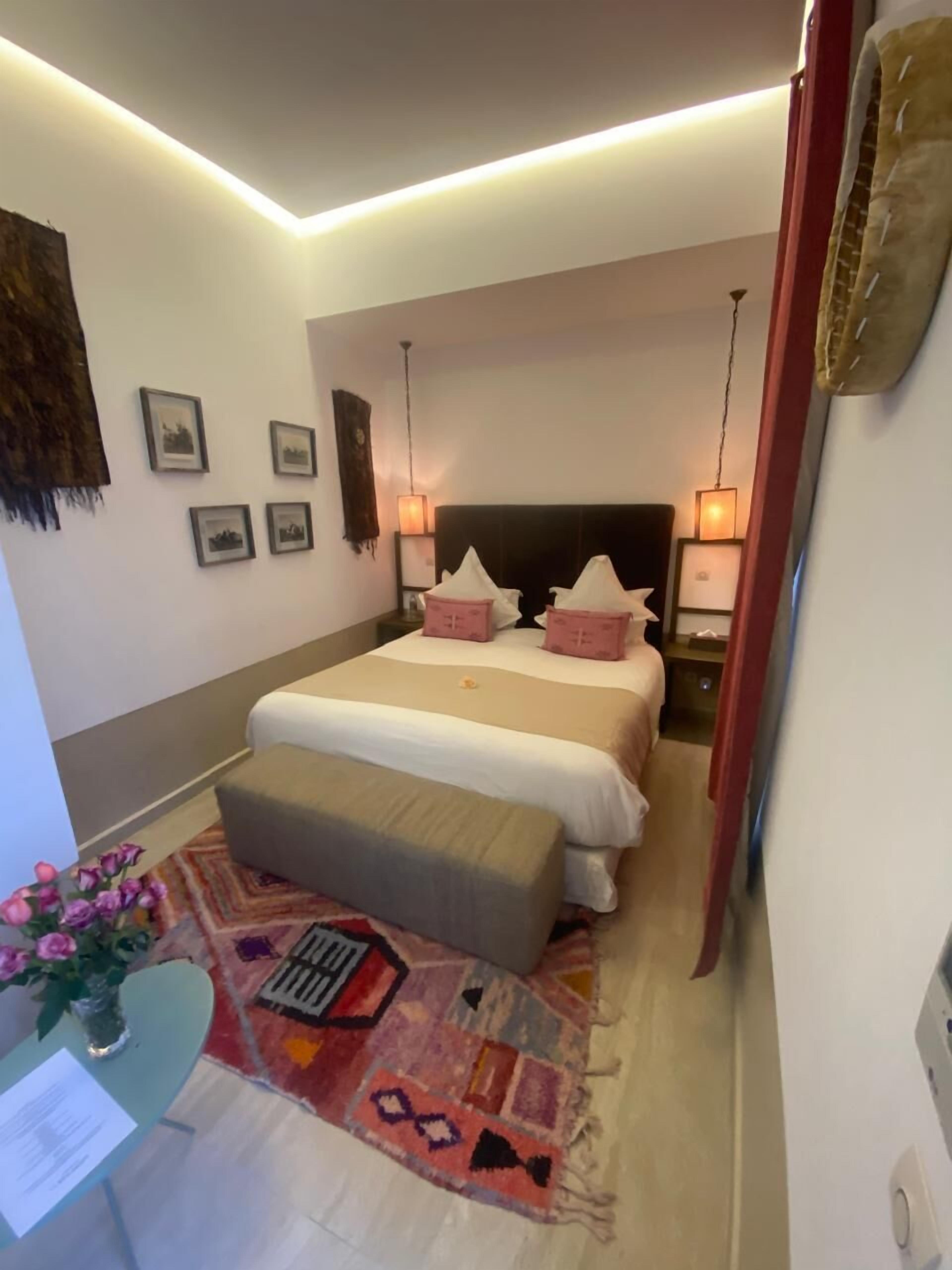 A hotel room with a bed, bedside tables, a desk with a chair, a rug, and framed pictures on the wall.