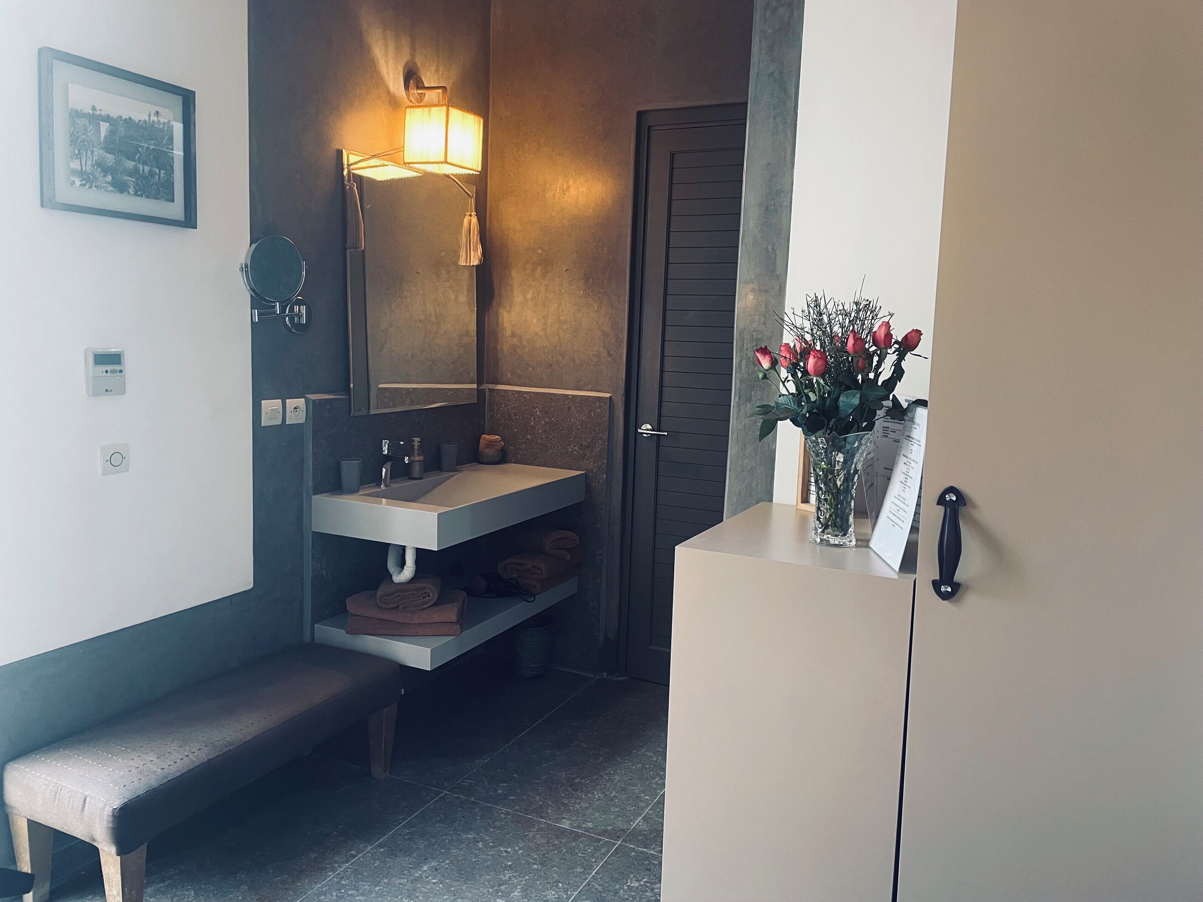 Une salle de bain équipée d’un lavabo, d’un miroir, d’un porte-serviettes, d’un banc et d’un vase avec des fleurs.