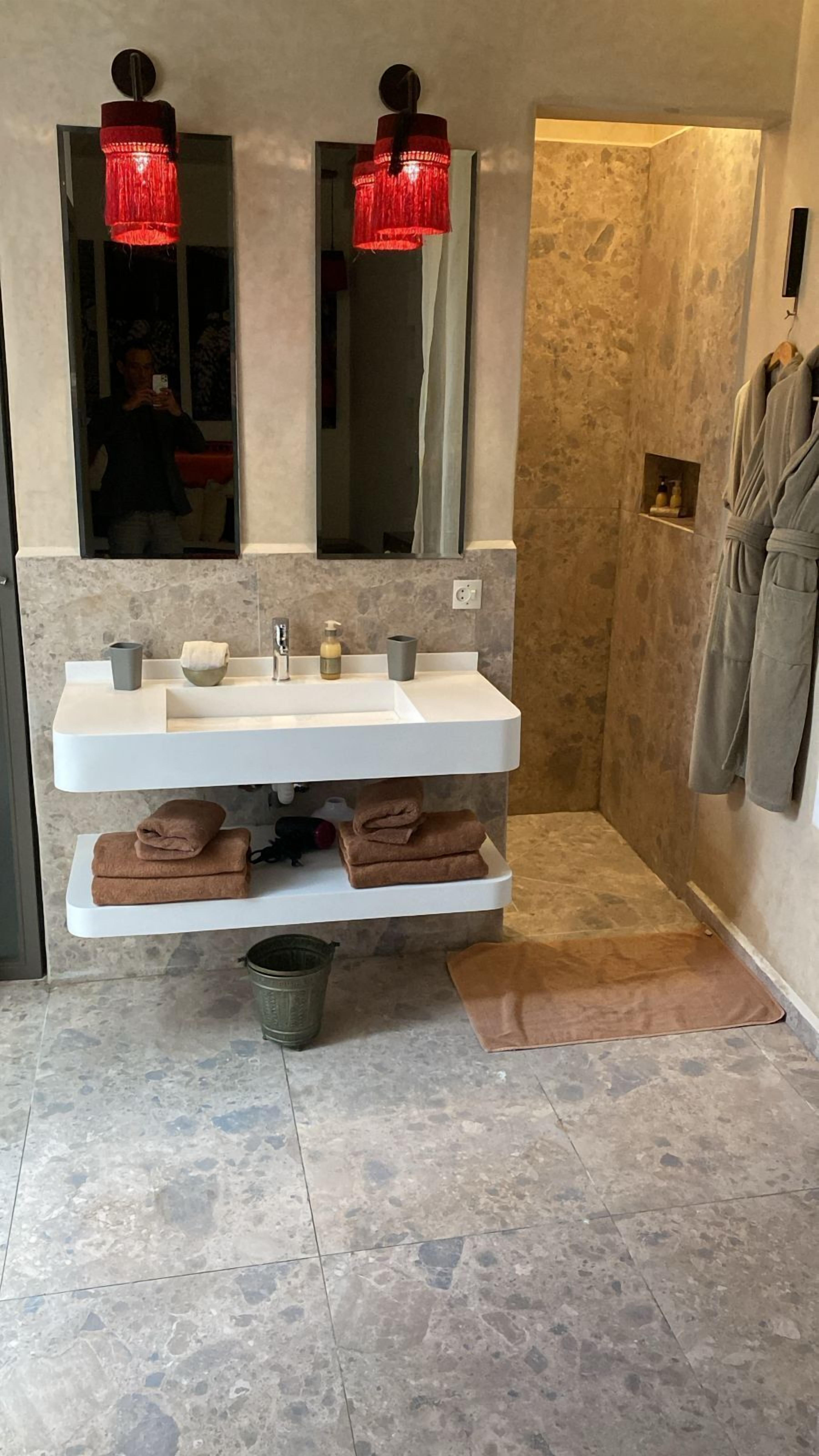 A bathroom with a white sink, two mirrors, red pendant lights, a towel rack, and a shower area.