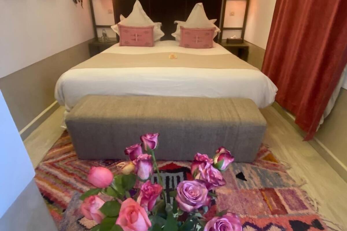 A bedroom with a bed, bedside tables, a nightstand, a bedside lamp, a window with curtains, and a vase with flowers.