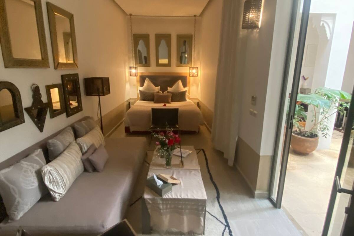 A modern living room with a sofa, a coffee table, and a bed with a headboard. There are wall-mounted lights and a large window.
