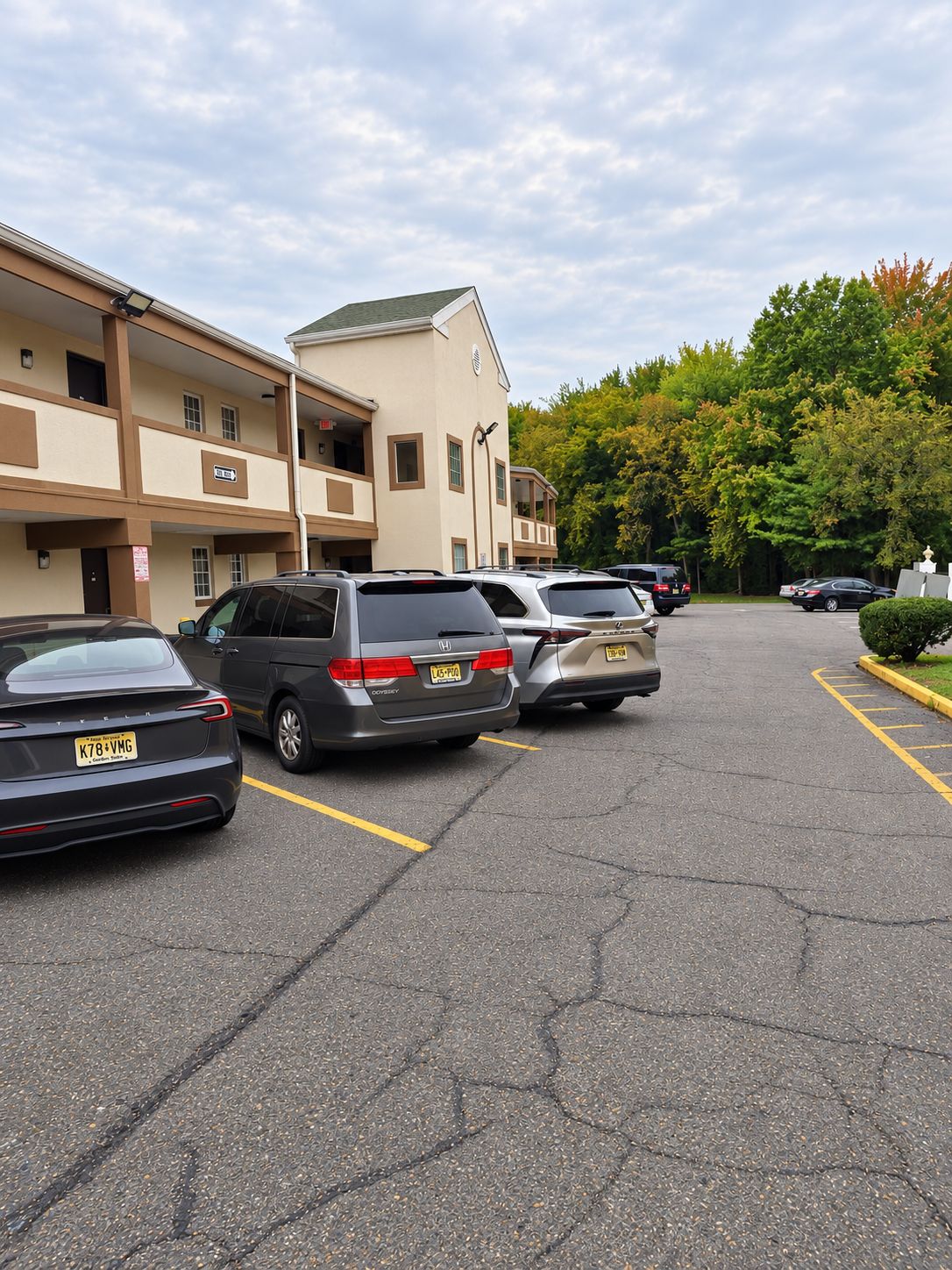 A parking lot with several cars, a building with multiple floors, and a green tree line in the background.