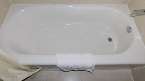 Une baignoire blanche standard, dotée d’un seul robinet et d’un drain, une serviette suspendue sur le côté et des murs recouverts de carreaux beiges.