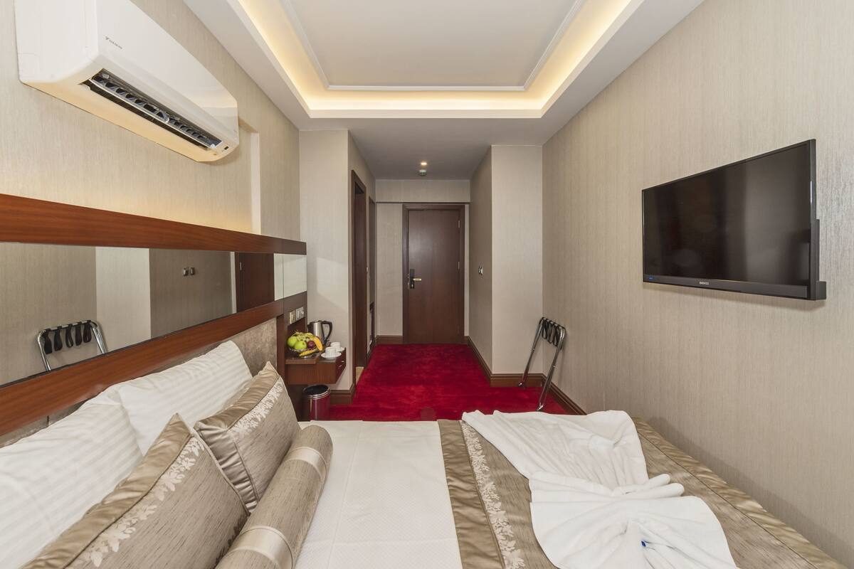 A hotel room with a bed, a TV mounted on the wall, a red carpet, and a hallway leading to a door.